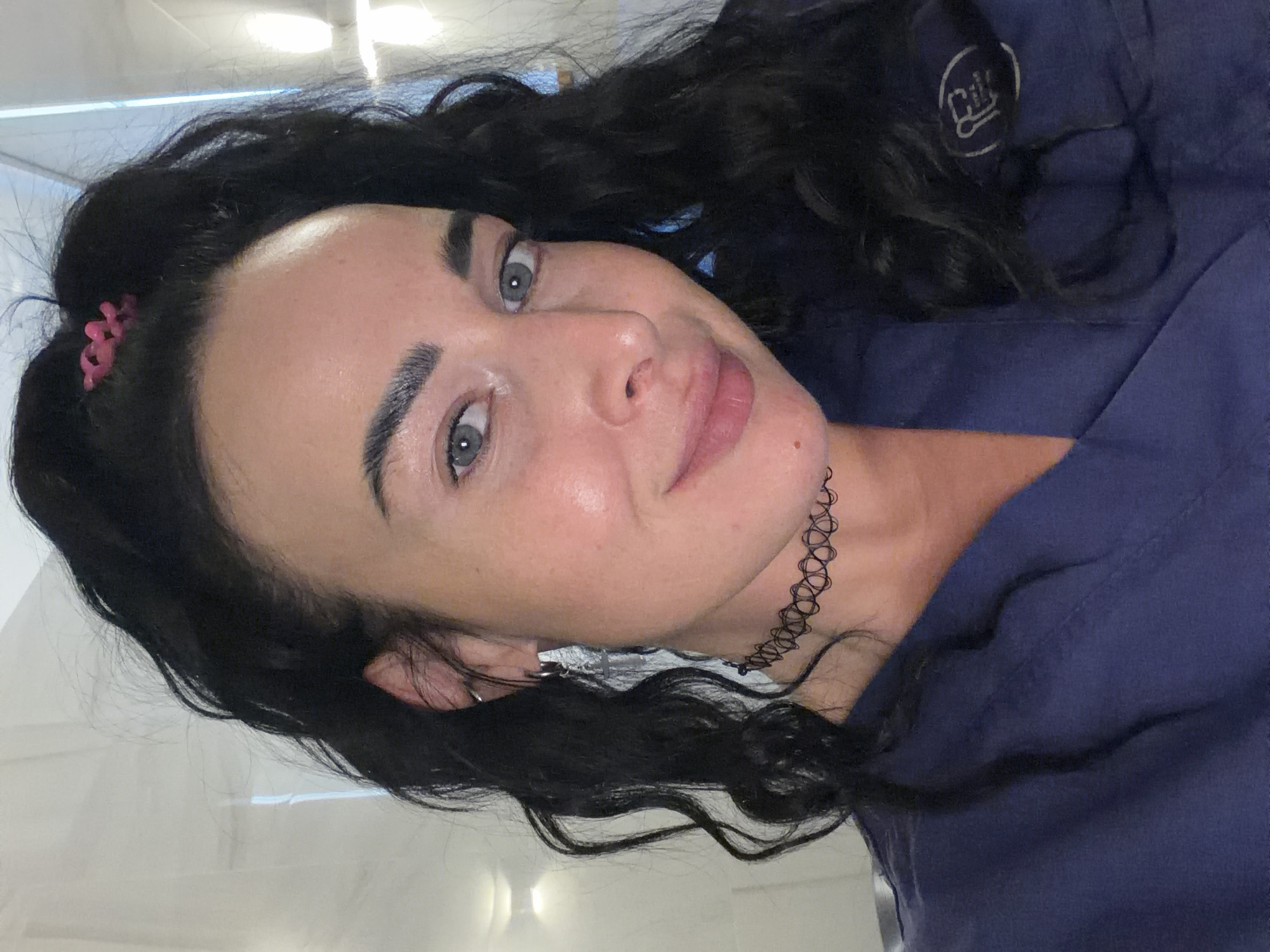 A woman with long, dark, curly hair and light eyes is the primary subject of the image. She is wearing a dark blue garment and a black choker necklace. A small pink hair clip is visible in her hair. The background appears to be an interior space with neutral colors. There is no evidence to suggest this image was taken in Saint Petersburg, Russia.