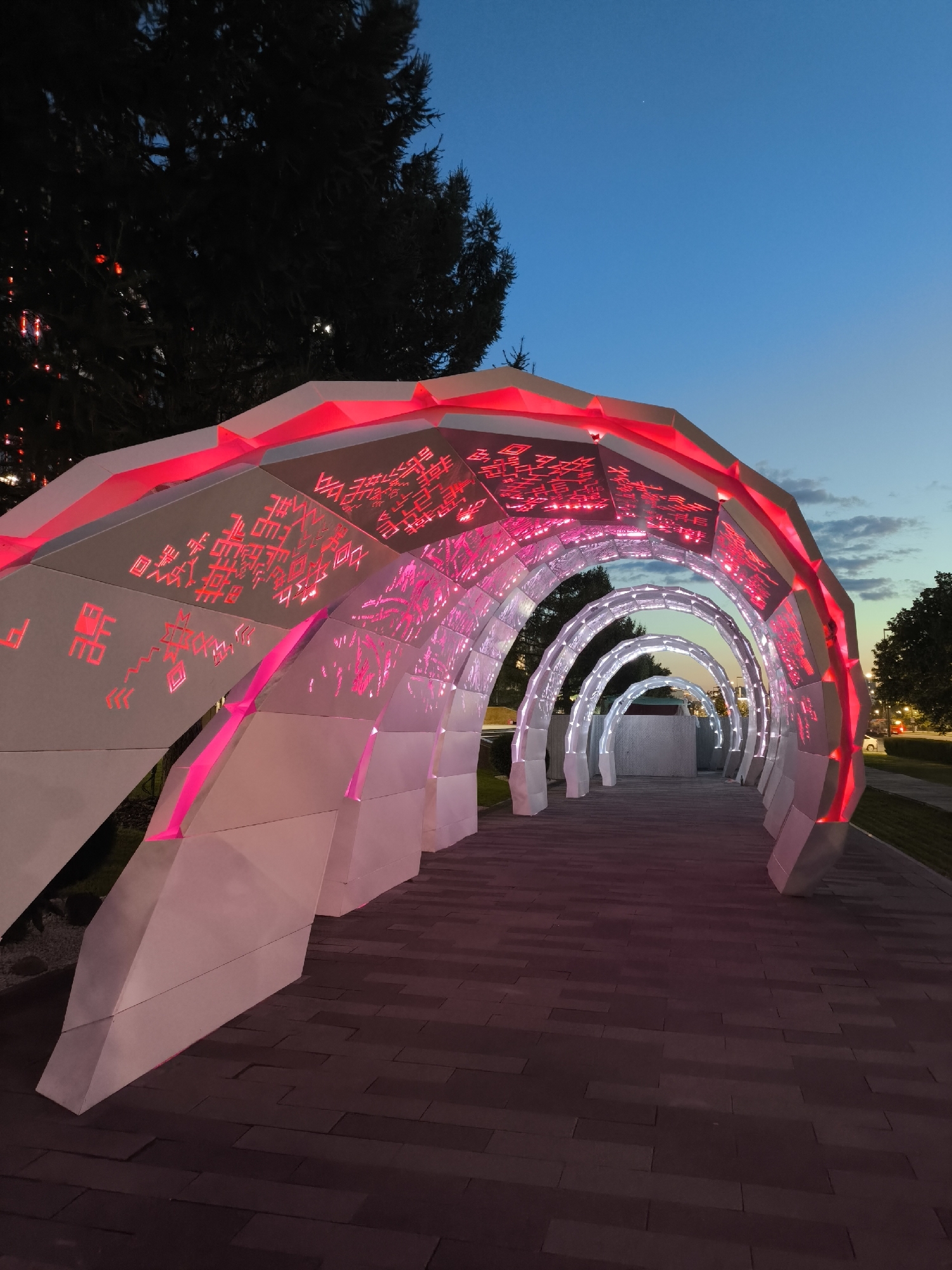 This is a low-angle, full shot of a modern, illuminated art installation at dusk in the city of Perm, Russia. The installation consists of a series of interconnected arched tunnels made of white, angular panels. The interior surfaces of the tunnels are patterned with red and white lights, creating a futuristic and artistic effect. The light emanates from within the structure, casting a pinkish glow on the surrounding paved walkway. The walkway is made of dark, rectangular paving stones laid in a patterned arrangement. To the left of the installation, the dark silhouette of a coniferous tree is visible against the twilight sky. The sky itself is a deep blue, transitioning to lighter shades near the horizon, with a few faint clouds. In the distance, through the arches of the installation, faint lights of urban elements and a streetlight are visible. There are no people or animals present in the frame. The overall scene conveys a sense of modern art and urban design in an outdoor public space at twilight.