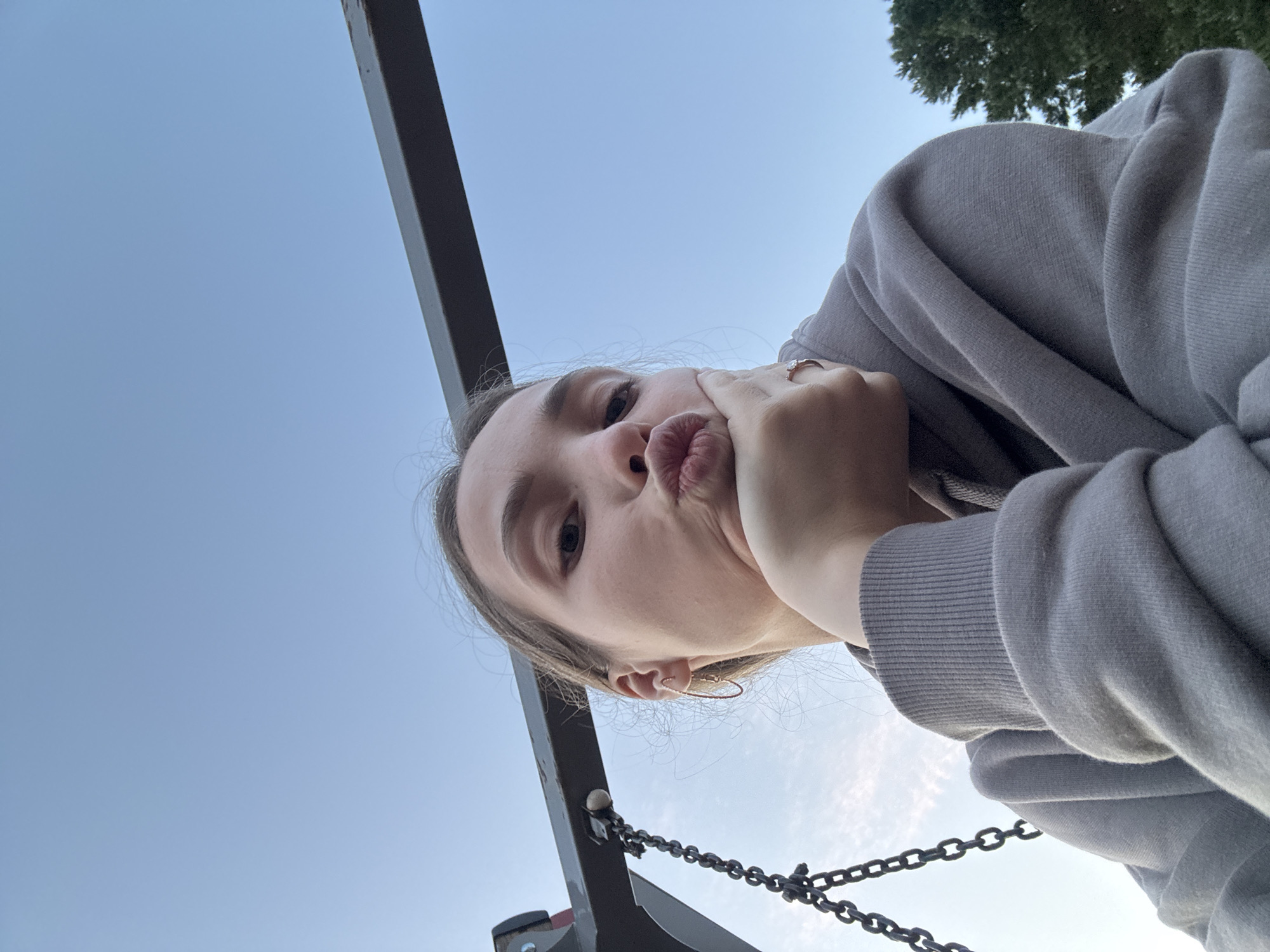 A young woman with her hair pulled back is taking a selfie outdoors. She is wearing a grey hoodie and has her hand on her chin. Her lips are puckered. In the background, the sky is blue with some faint clouds. A metal bar and a chain are visible in the upper left of the frame, suggesting the presence of playground equipment like a swing set. A tree is visible on the right side of the frame. The location is not specified as Noviy, Russia.