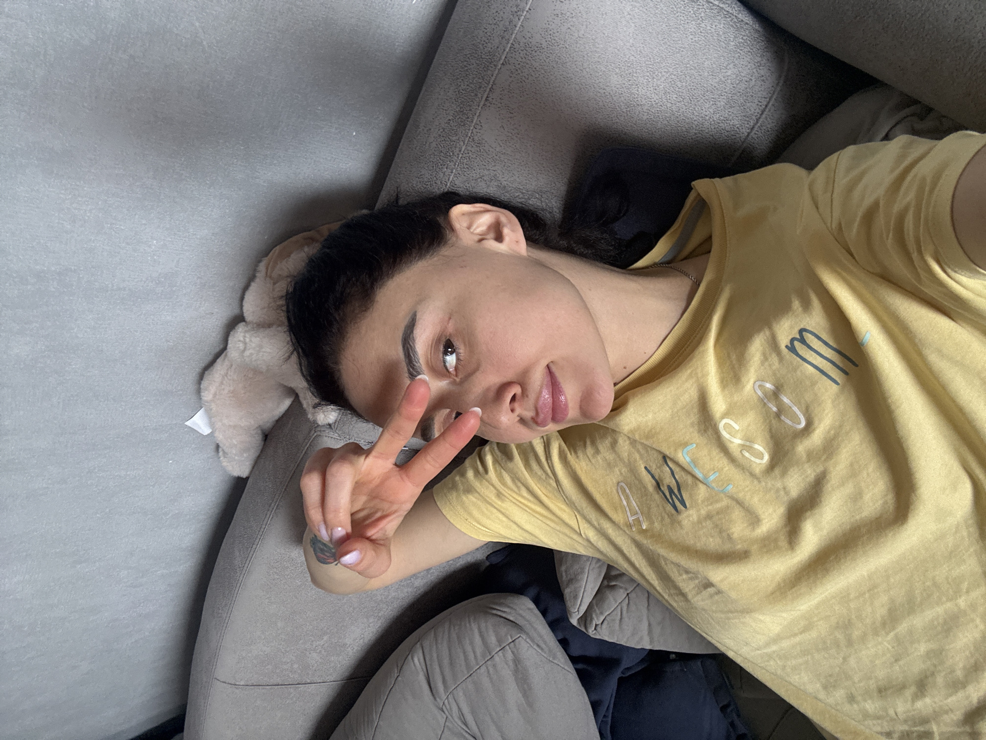 A woman lies on a gray couch. She wears a yellow t-shirt with the word "Awesome" on it. She is making a peace sign with her left hand. The background includes a beige stuffed animal near her head and what appears to be clothing beneath her. The location cannot be determined to be the city of Tbilisi, Georgia from this image.