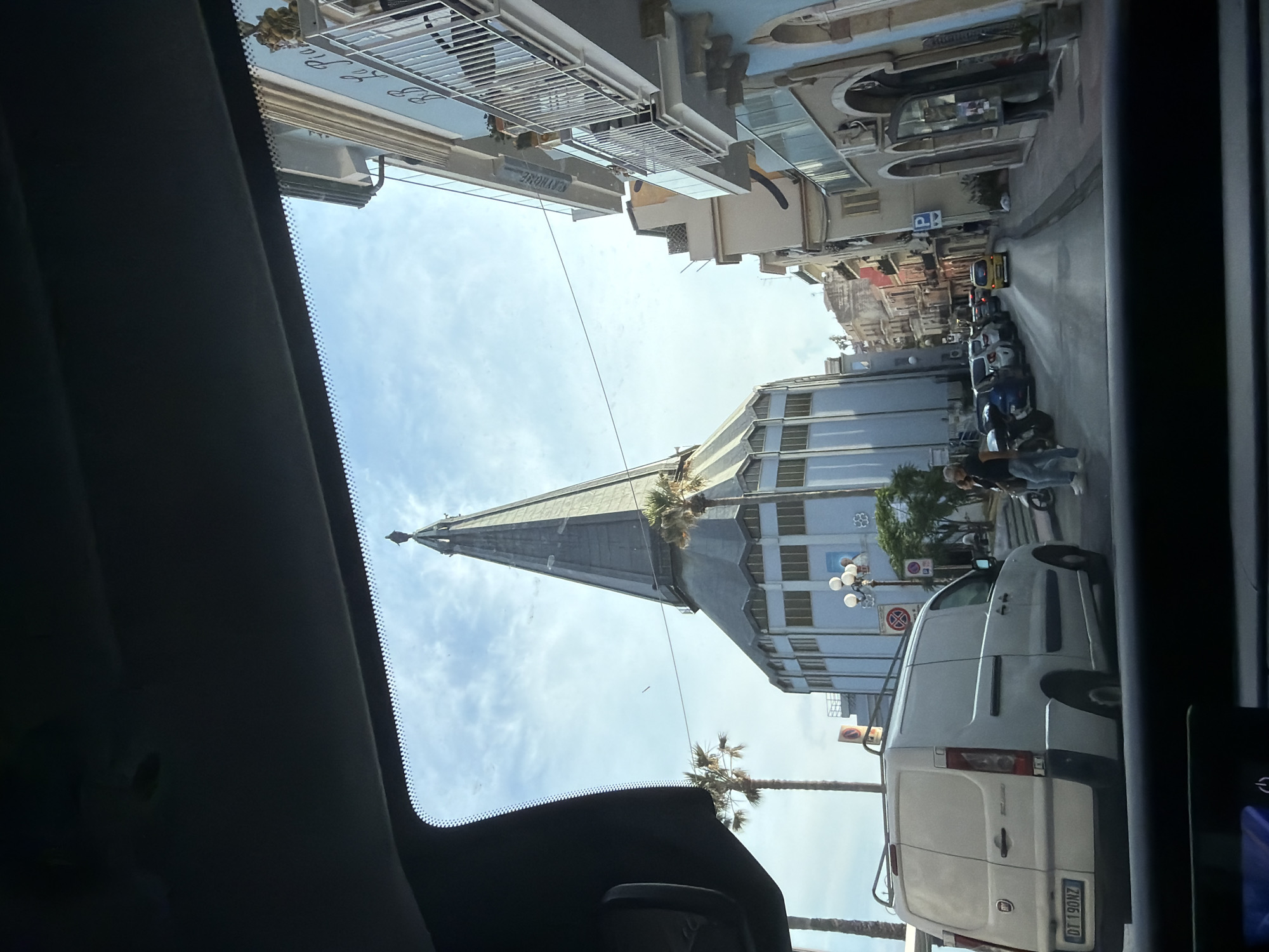 A view from inside a vehicle shows the Basilica of St. Peter and Paul towering above the street in Chianchitta, Italy. The basilica's gray spire dominates the skyline. The street scene includes parked motorcycles and a white Fiat van. Pedestrians are present near the vehicles. Buildings line the street on either side. The sky is light blue with scattered clouds. The vehicle's interior elements are visible in the foreground.