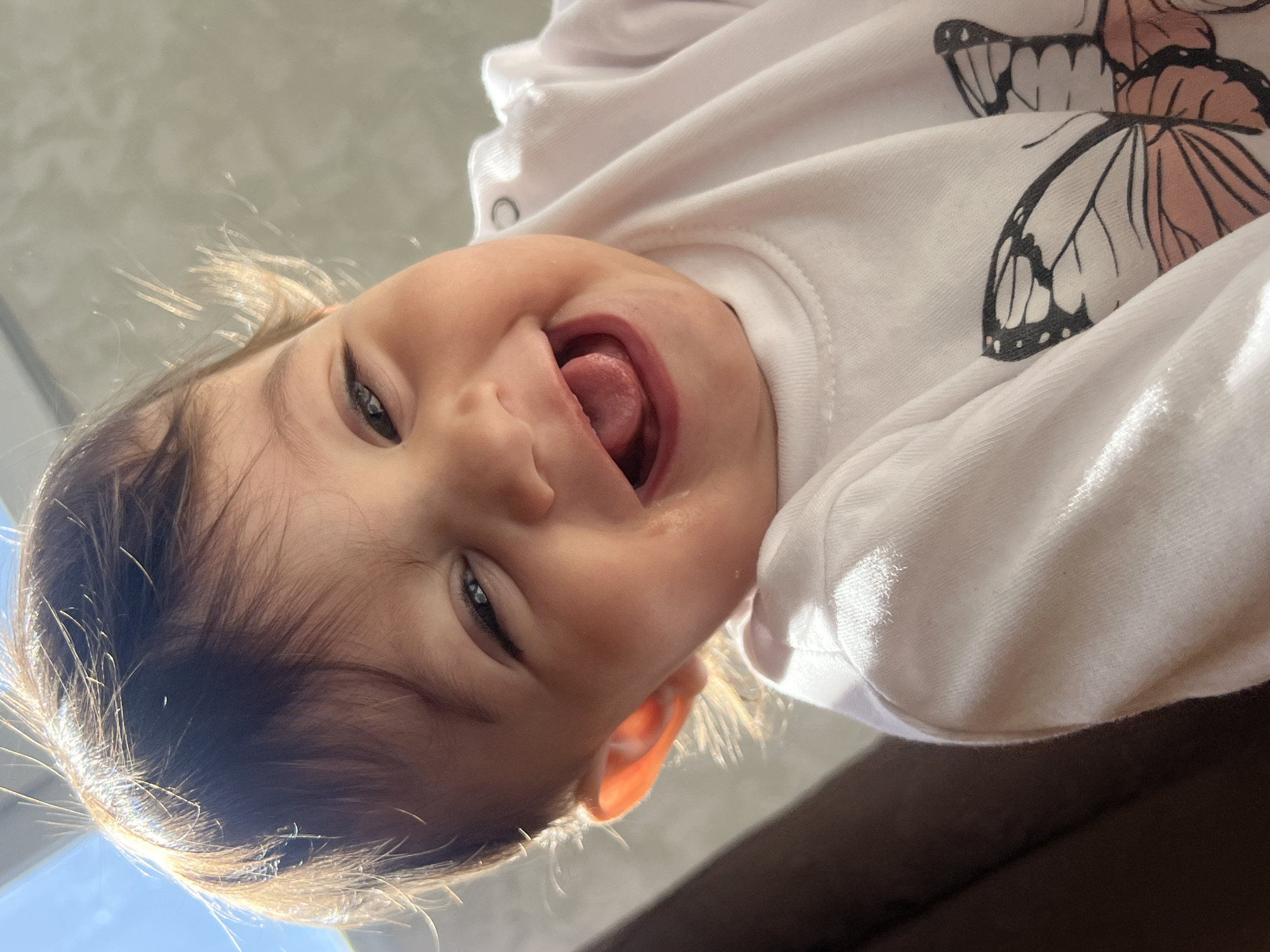 A baby wearing a white shirt with a butterfly print is looking down at the camera with their mouth open and tongue sticking out. The image is taken from a low angle, looking up at the baby. Light streams in from the top left, illuminating the baby's hair. The background consists of a neutral-colored wall or surface in the top left, and a dark surface in the bottom right. The location is not discernible from the image.