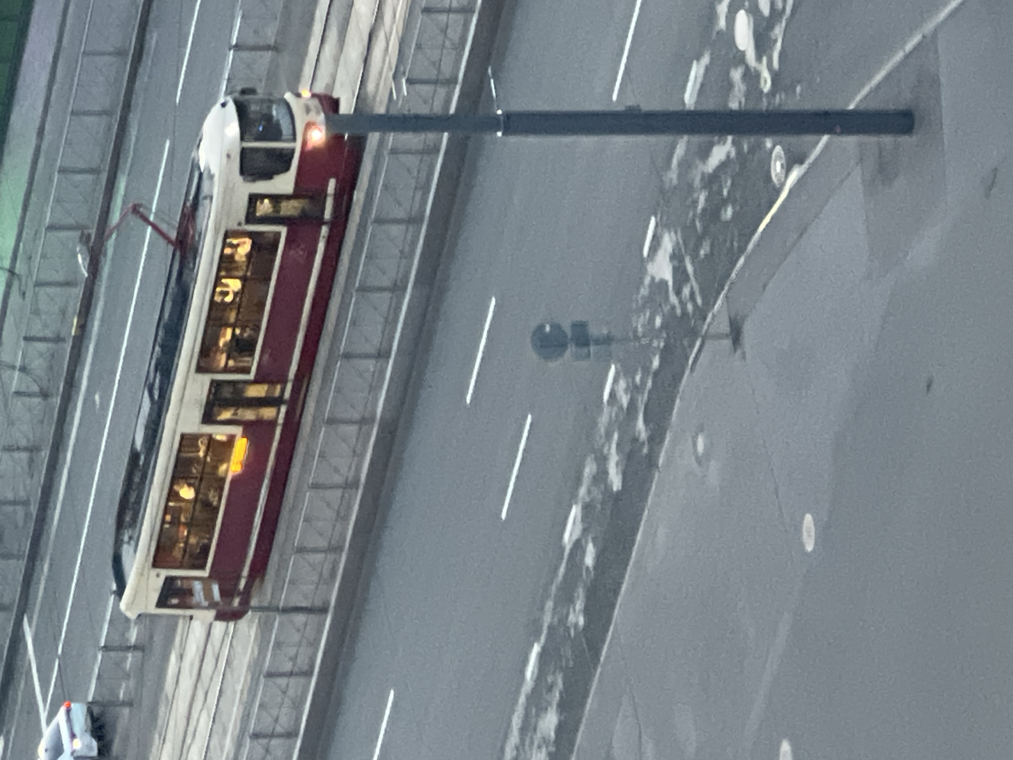 A high-angle shot captures a red and white tram with illuminated windows moving along tram tracks next to a road. A white car is visible on the road to the left of the tram. The environment appears to be an urban setting with a paved road and sidewalks. A lamppost stands on the right side of the frame, and a traffic sign is visible on the left. The lighting suggests it is either dusk or a cloudy day.