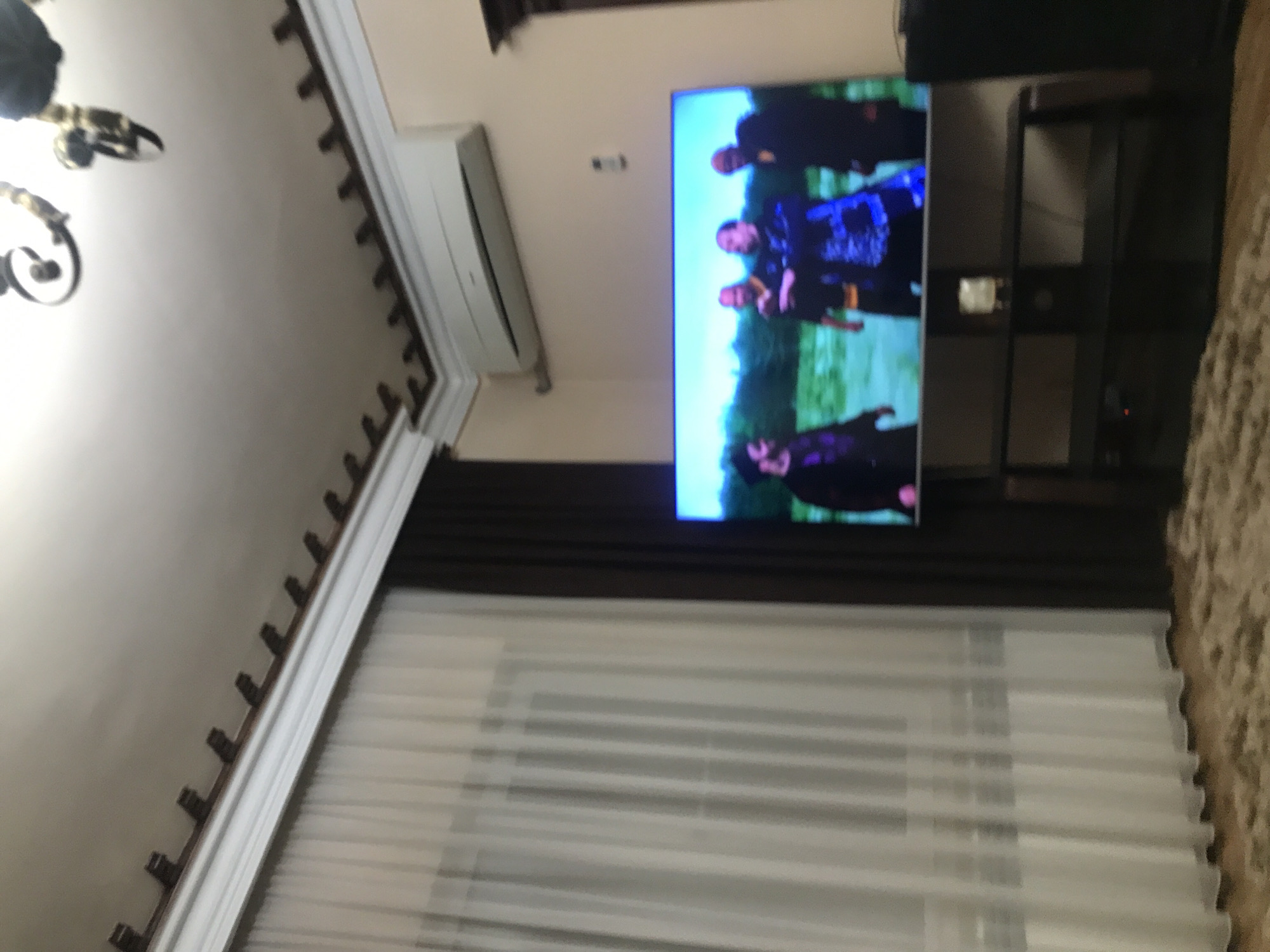 The image depicts an interior scene, possibly within a residential space. A television screen displays what appears to be a group of people on screen. Below the television, there is a dark-colored entertainment unit. To the left of the television, a curtain hangs, with a section of translucent white fabric covering a window. Above the television, there is an air conditioning unit mounted on the wall. The ceiling has decorative molding and a chandelier can be seen in the upper left. The overall environment suggests a living room or entertainment area. The location within Yerevan, Armenia cannot be verified.
