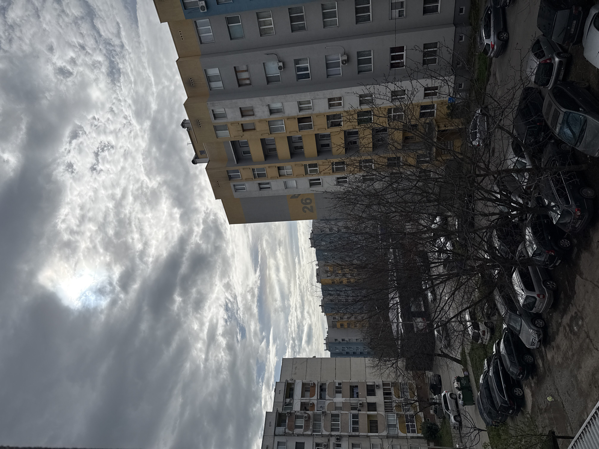 The image depicts an overcast urban landscape, likely taken from an elevated position. Several multi-story apartment buildings are visible, with a consistent style indicative of post-World War II Soviet-era architecture. The building closest to the viewer is grey and yellow, with the number "26" painted on its side. A row of bare trees lines the street below, partially obscuring a parking area filled with cars. The sky is filled with heavy, grey clouds. The location is Stara Zagora, Bulgaria.