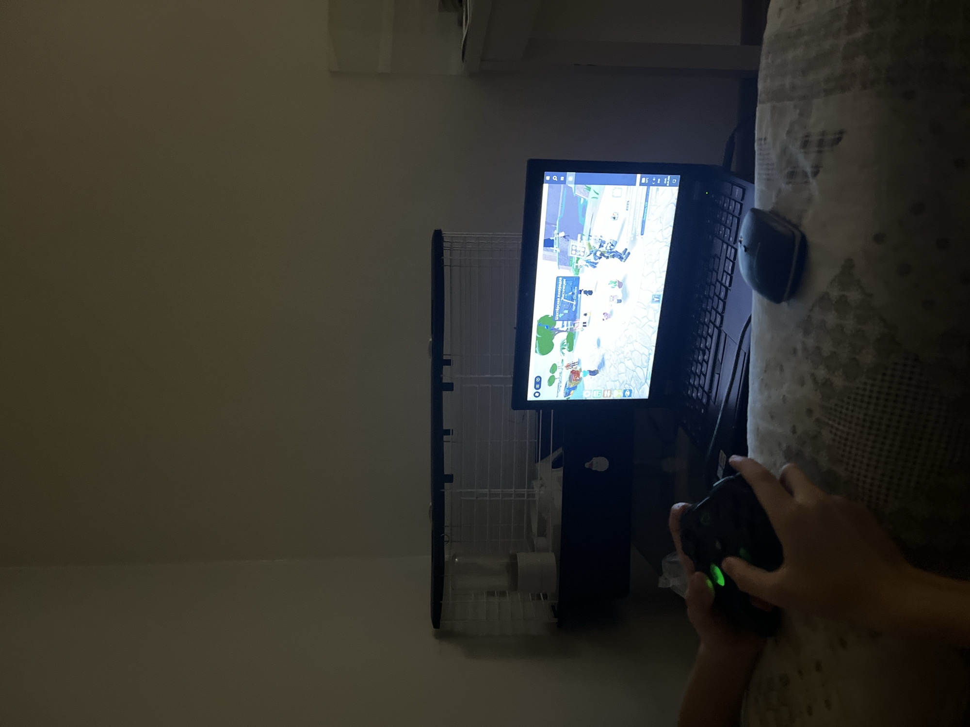 A person is playing a video game on a monitor. The monitor is resting on a small stand, which is next to a bed. The person is holding a controller in their hands, and the game appears to be a battle royale. The environment appears to be a room indoors with a blank wall and overhead lighting. The location in the city of Unknown, Unknown is indeterminable.