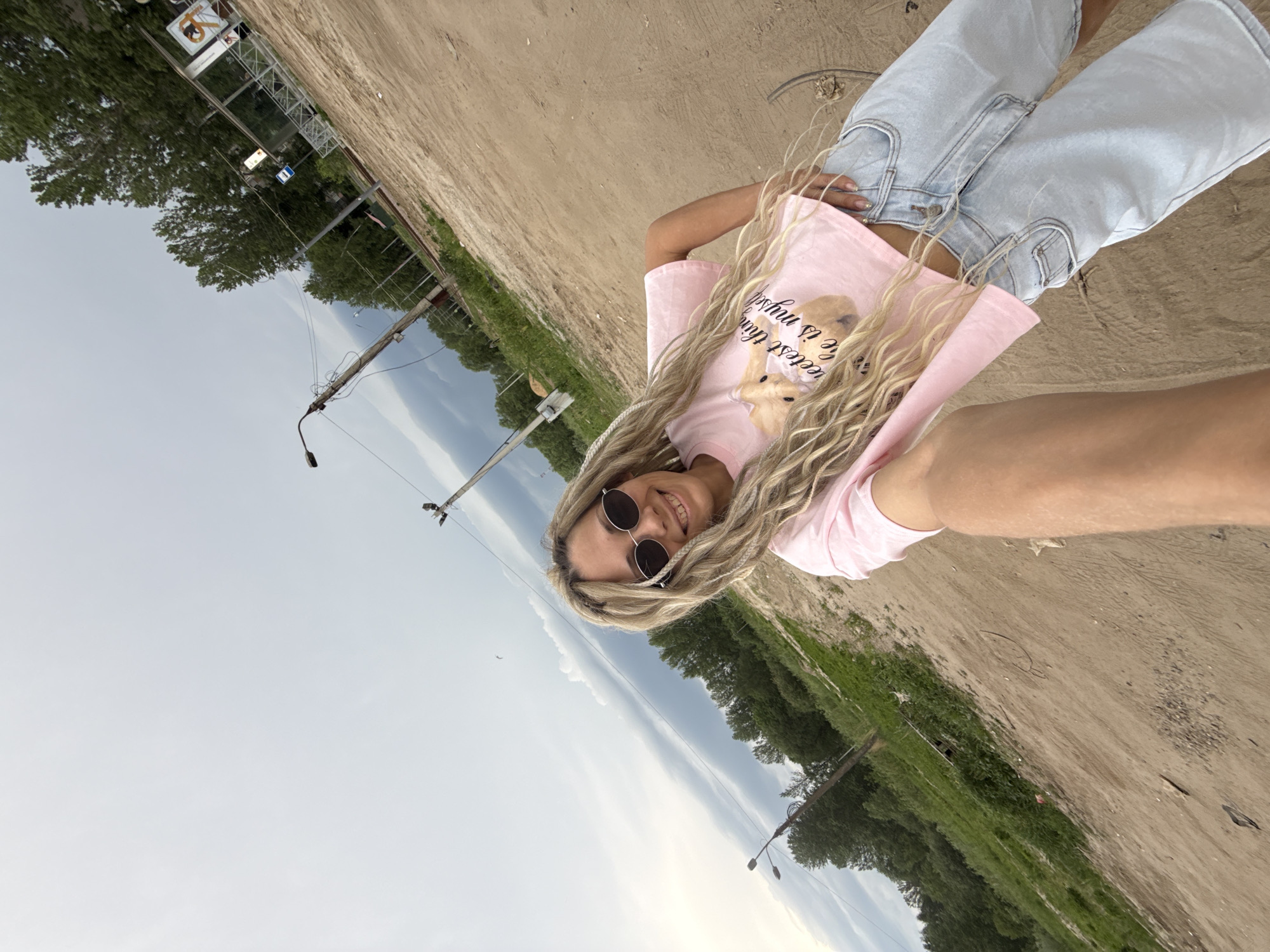 A young woman with blonde dreadlocks, sunglasses, and a pink T-shirt stands on a sandy surface in Rusanovo, Russia. She is wearing light blue jeans. The scene includes trees, grass, a cloudy sky, and utility poles with lights.