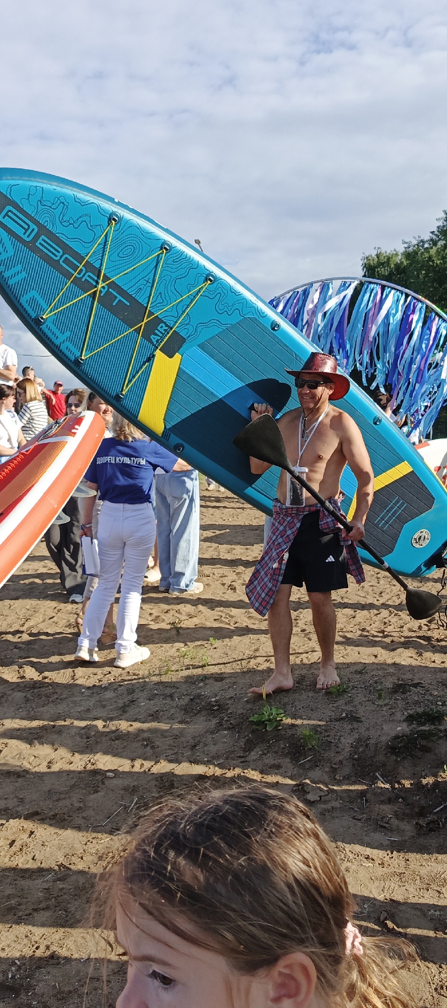 In Chaikovsky, Russia, at what appears to be a waterfront event or festival, a man wearing a red cowboy hat, sunglasses, and a plaid shirt tied around his waist is carrying a large blue and yellow paddleboard. He is barefoot and holding a paddle. Behind him, a woman in white pants and a blue t-shirt with "ДВОРЕЦ КУЛЬТУРЫ" (Palace of Culture) printed on it stands holding a white object. Other people are visible in the background, along with decorative blue and purple streamers hanging from a structure. The ground is sandy and there is some vegetation. A portion of a child's head is in the foreground, on the right side of the image.