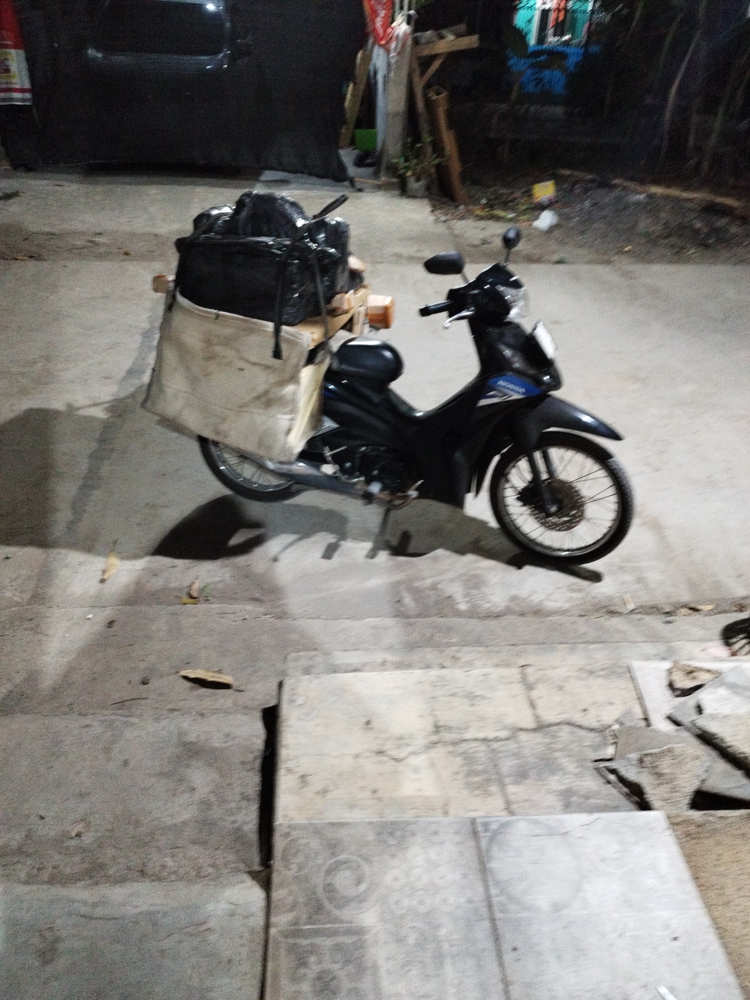 A black motorcycle is parked on a concrete surface at night. The motorcycle has a large, light-colored canvas bag strapped to the back, filled with black, plastic-wrapped bundles. The motorcycle has a visible license plate, though it is not legible. In the background, a dark-colored vehicle is partially visible, along with some wooden structures and foliage. The scene appears to be an outdoor area, possibly a street or alleyway, in Karawang, Indonesia, given the context provided. The lighting suggests it is evening or night.
