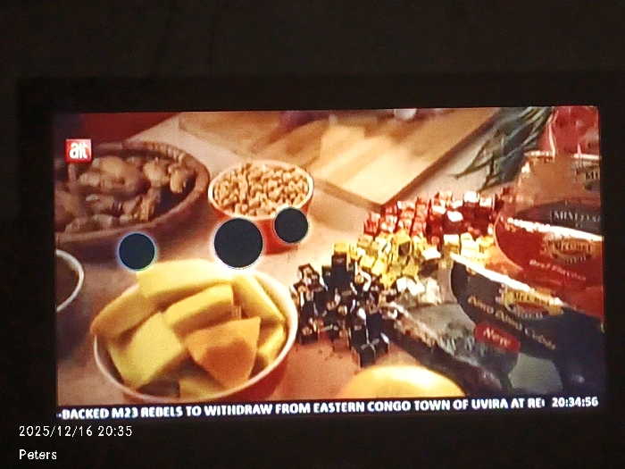 A television screen displays various food items in bowls and packages. There's a bowl of meat, a bowl of beans, and a bowl filled with what appear to be cubed mangoes and sweet potatoes. Packages of mixed diced beef flank the cubed ingredients, which are arranged in piles. Text at the bottom of the screen reads, "BACKED M23 REBELS TO WITHDRAW FROM EASTERN CONGO TOWN OF UVIRA AT REI 20:34:56 2025/12/16 20:35 Peters" alongside the "alt" logo.