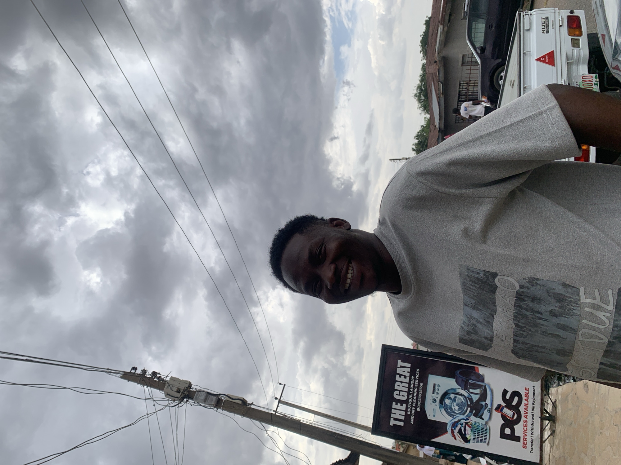 The image features a smiling young man in an outdoor urban setting, likely a street or roadside in Jos, Nigeria, under an overcast sky. The photograph is taken from a slightly low angle, looking up towards the clouds.
The main subject is a person with dark skin and short dark hair, wearing a light grey t-shirt with a faded graphic design that partially shows text like "DUE" and "U-". He is positioned to the right of the frame, looking directly at the camera with a broad, genuine smile, revealing his teeth and conveying a cheerful mood.
In the background, to the left, a multi-wired electrical pole with various distribution boxes is prominent, typical of street infrastructure. Below the pole and further back, a black advertising board stands out. The board promotes "THE GREAT BOUTIQUE, LAUNDRY AND DRY CLEANING SERVICES" along with the phone number "08036938552". It also advertises "POS SERVICES AVAILABLE" for "Transfer | Withdrawal | Bill Payment".
Further behind the subject and the board, a street scene unfolds with vehicles and buildings. A white pickup truck, bearing "HIJET DAIHATSU" branding and a "C CAUTION" sticker, is visible, with a partial license plate "JOXD". Another dark vehicle is partially seen beside it. The buildings appear to be single or two-story structures with corrugated roofs. The sky is largely covered with thick, grey clouds, suggesting an overcast day.