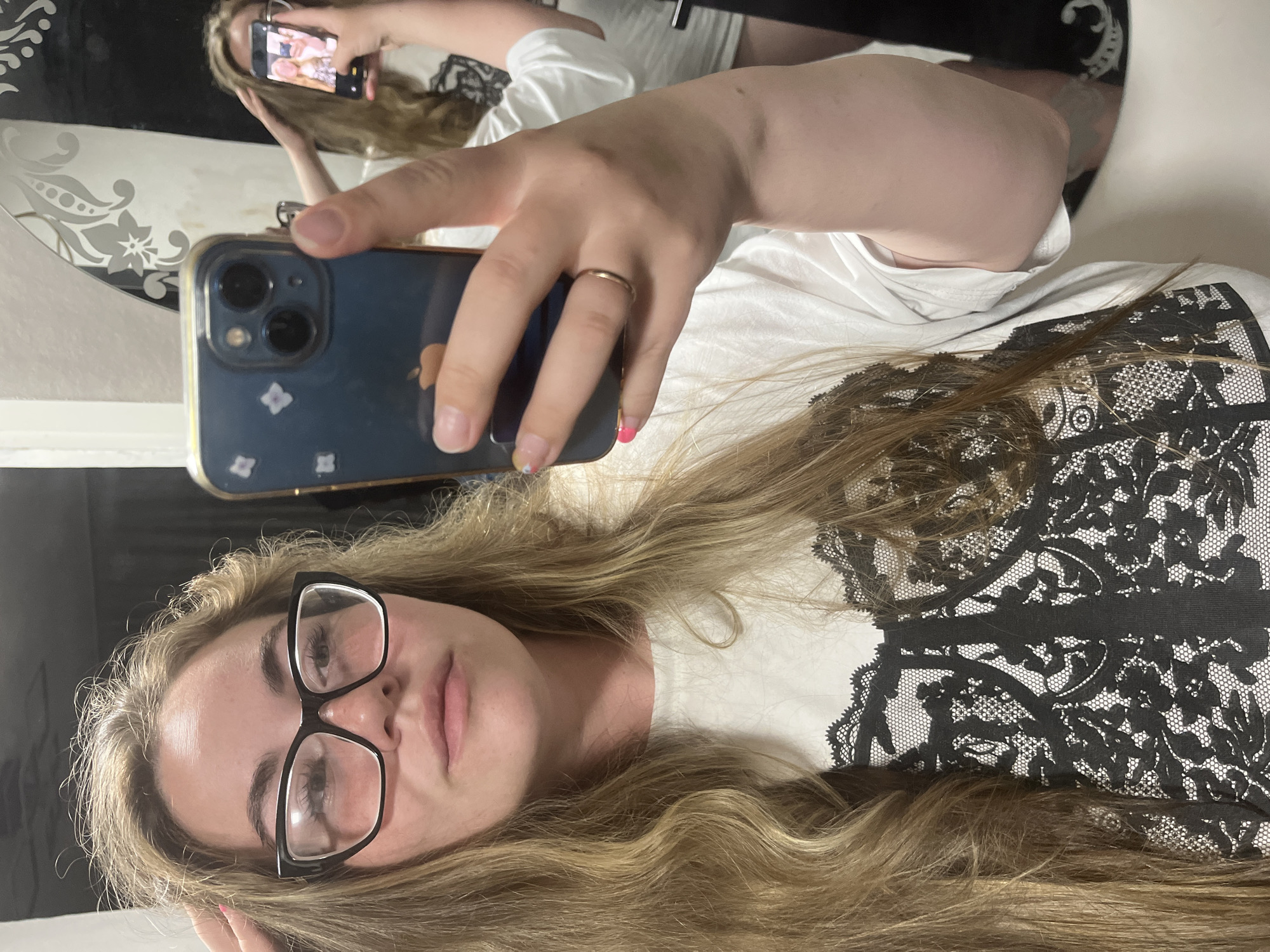 A young woman with long blonde hair and glasses is taking a selfie in a bathroom. She is wearing a white t-shirt with a black lace design. She is holding a smartphone in her right hand. In the background, a mirror reflects another person, presumably the same woman, also taking a selfie. The bathroom appears to be modern, with white tiles and a patterned wallpaper. The location is identified as Chaikovsky, Russia.