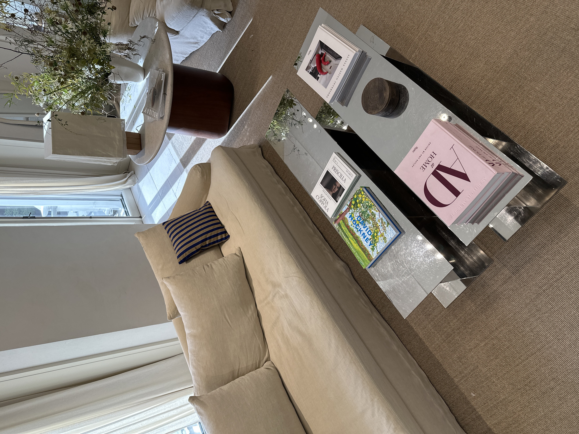 This is an interior shot of a living room. A beige sofa with several pillows, including one with blue stripes, is the focal point. Next to the sofa is a set of nested mirrored coffee tables. The tables are decorated with books, including titles such as "Priscilla Sofia Coppola," "David Hockney Fondation Louis Vuitton," "AD at HOME," and "ALESSA KAMITO INTERIORS." A small wooden container also sits on the table. To the left of the sofa is a round side table, partially obscured, with a vase of flowers on top. The room has beige carpet, and white curtains can be seen near a window in the lower-left corner. The image does not provide any information to determine whether the location is in Madrid, Spain.