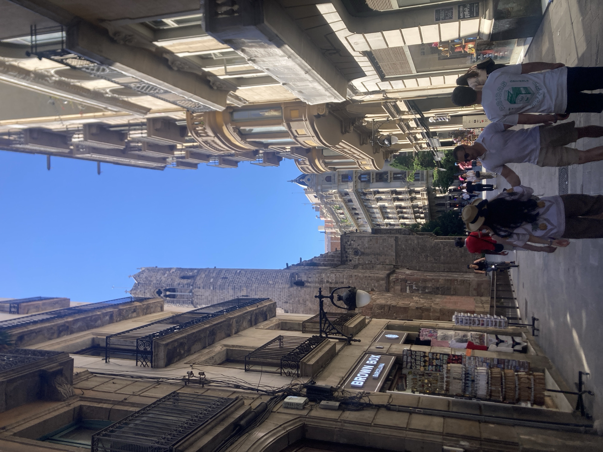 The image shows a street scene in Barcelona, Spain, under a bright blue sky. The perspective is looking upward at the buildings lining the street. On the left side, the buildings are older with ornate balconies and detailed facades, featuring a sign for "BROWN BOX BARCELONA." On the right, there's a tall stone structure with a tower, likely part of a church or historical building. The street is paved and crowded with pedestrians. Some people are walking, and some are standing near merchandise displays. The buildings on the opposite side of the street are taller with modern architectural elements. Several people walking in the background.