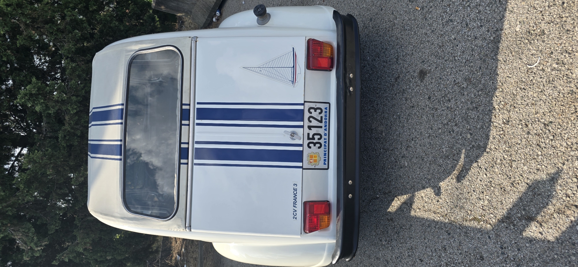 The image shows the rear of a white Citroën 2CV with blue racing stripes. The car has a license plate from Andorra that reads "35123 PRINCIPAT D'ANDORRA". To the left of the license plate, the text "2CV FRANCE 3" is visible. On the right side of the rear of the car, there is a sticker of a sailboat. The car is parked on an asphalt surface, with shadows cast on the ground. In the background, there are trees and a concrete barrier. There is no indication of the city of Collbató, Spain in the image.