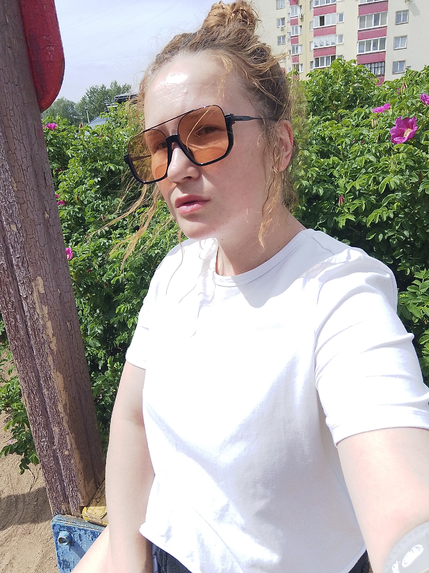 A fair-skinned person with reddish-blonde hair pulled into a messy bun is taking a selfie. They are wearing large, square sunglasses with orange lenses and a white t-shirt. The background consists of dense green bushes with pink flowers, and a multi-story apartment building with pink and white trim is visible in the upper right. The person appears to be sitting on a wooden structure, possibly part of a playground. The location is the city of Chaikovsky, Russia.