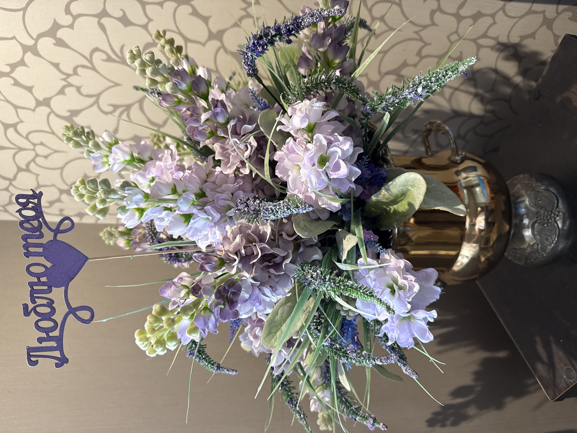 A vibrant bouquet of artificial flowers, primarily in shades of light purple, lavender, and white, with green foliage and buds, is the central subject of this indoor photograph. The flowers are arranged in a golden, metallic vase or urn, featuring ornate handles, one of which is partially visible on the right.
The bouquet rests on a smooth, light-colored surface, likely a table, which reflects some of the light. The background is a light beige or off-white wall adorned with a repeating, scrolling pattern that casts subtle shadows from the flowers. The soft, ambient lighting suggests an indoor setting, possibly a home, in Votkinsk, Russia.
A notable detail is a decorative purple cutout placed on the left side of the bouquet. This cutout spells out the Russian phrase "Люблю тебя" in a script font, accompanied by a heart shape, which translates to "I love you" in English. This element suggests a theme of affection or celebration. The image captures a still life, showcasing a decorative arrangement.