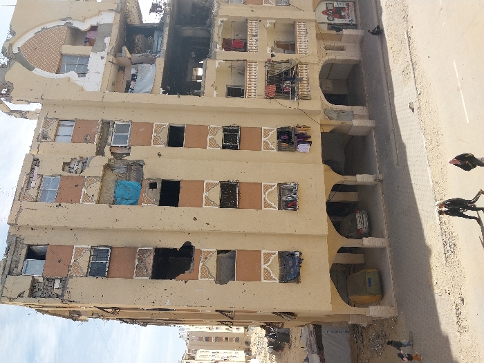 A multi-story building exhibits extensive damage, likely from conflict or disaster. Windows are broken or missing, and structural supports are visibly compromised. There are gaping holes in the facade. Possessions like clothes hang from windows. People can be seen in the street near the building. The building’s lower level has carports, and vehicles are parked within them. The scene conveys a sense of destruction and displacement.