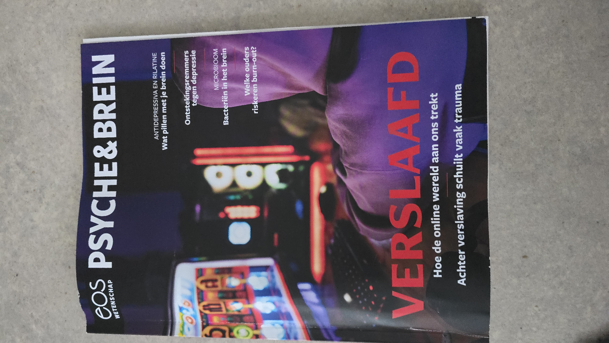 The image shows a magazine called "Eos PSYCHE&BREIN" with the headline "VERSLAAFD" in large, red letters, meaning "addicted." Smaller text reads "Hoe de online wereld aan ons trekt" ("How the online world attracts us") and "Achter verslaving schuilt vaak trauma" ("Behind addiction often lies trauma"). There are also smaller articles listed, such as "Antidepressiva en Rilatine: Wat pillen met je brein doen" ("Antidepressants and Ritalin: What pills do with your brain"), "Ontstekingsremmers tegen depressie" ("Anti-inflammatory drugs against depression"), "Microbioom: Bacteriën in het brein" ("Microbiome: Bacteria in the brain"), and "Welke ouders riskeren burn-out?" ("Which parents risk burnout?"). The cover image features a person wearing a purple hoodie in front of a slot machine.
