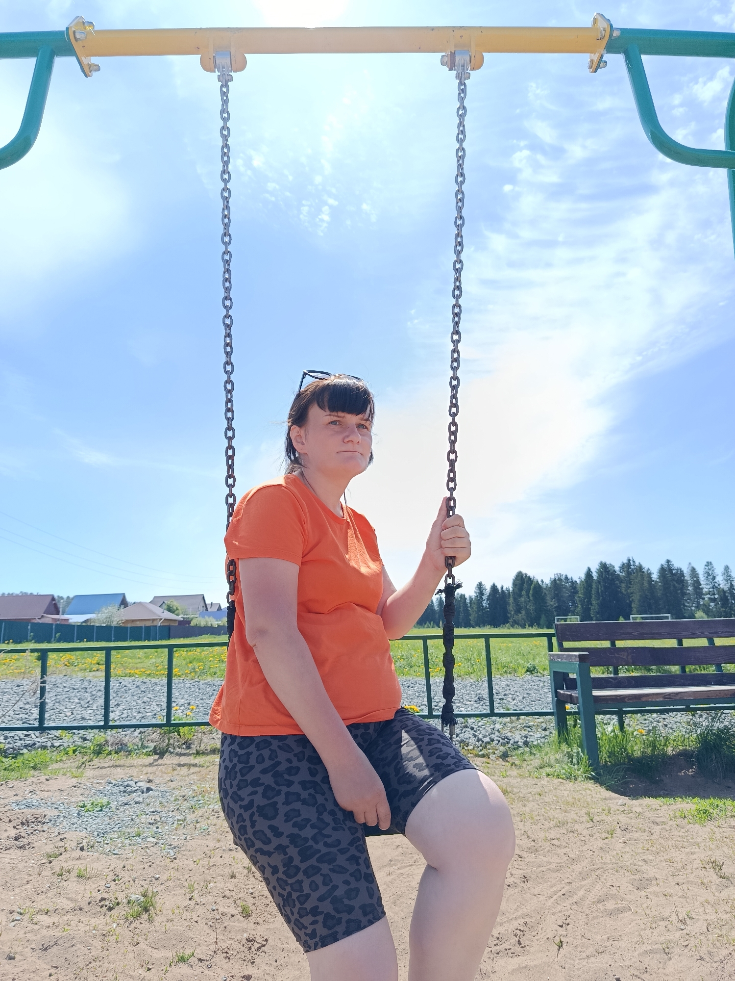 The image shows a woman on a swing in a playground. She is wearing an orange T-shirt and leopard print shorts. She is holding the swing chain with one hand. The playground has a yellow and green swing set. In the background, there is a green fence, a bench, grass, and some houses. The sky is blue with some clouds. The location is Фоки, Russia.