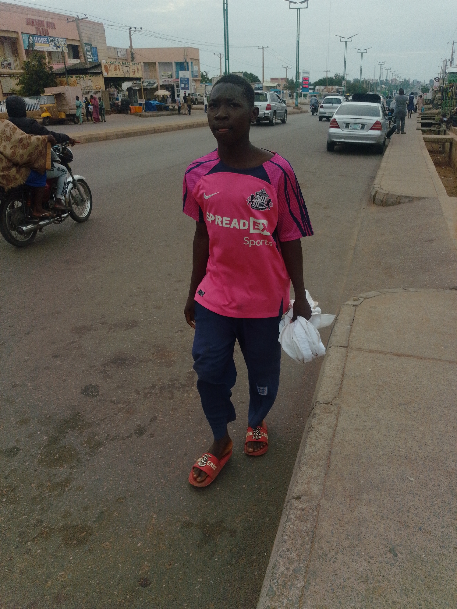 The image shows a young Black male walking on the side of a street in Gombe, Nigeria. He is wearing a bright pink t-shirt with blue trim and dark blue pants. He is also wearing red sandals. In his right hand, he is carrying a white bundle of fabric. The street is paved and appears to be a commercial area, with buildings lining one side. There are several vehicles on the street, including cars and a motorcycle. The sky is overcast. The area is a busy urban environment with typical street-level commerce.