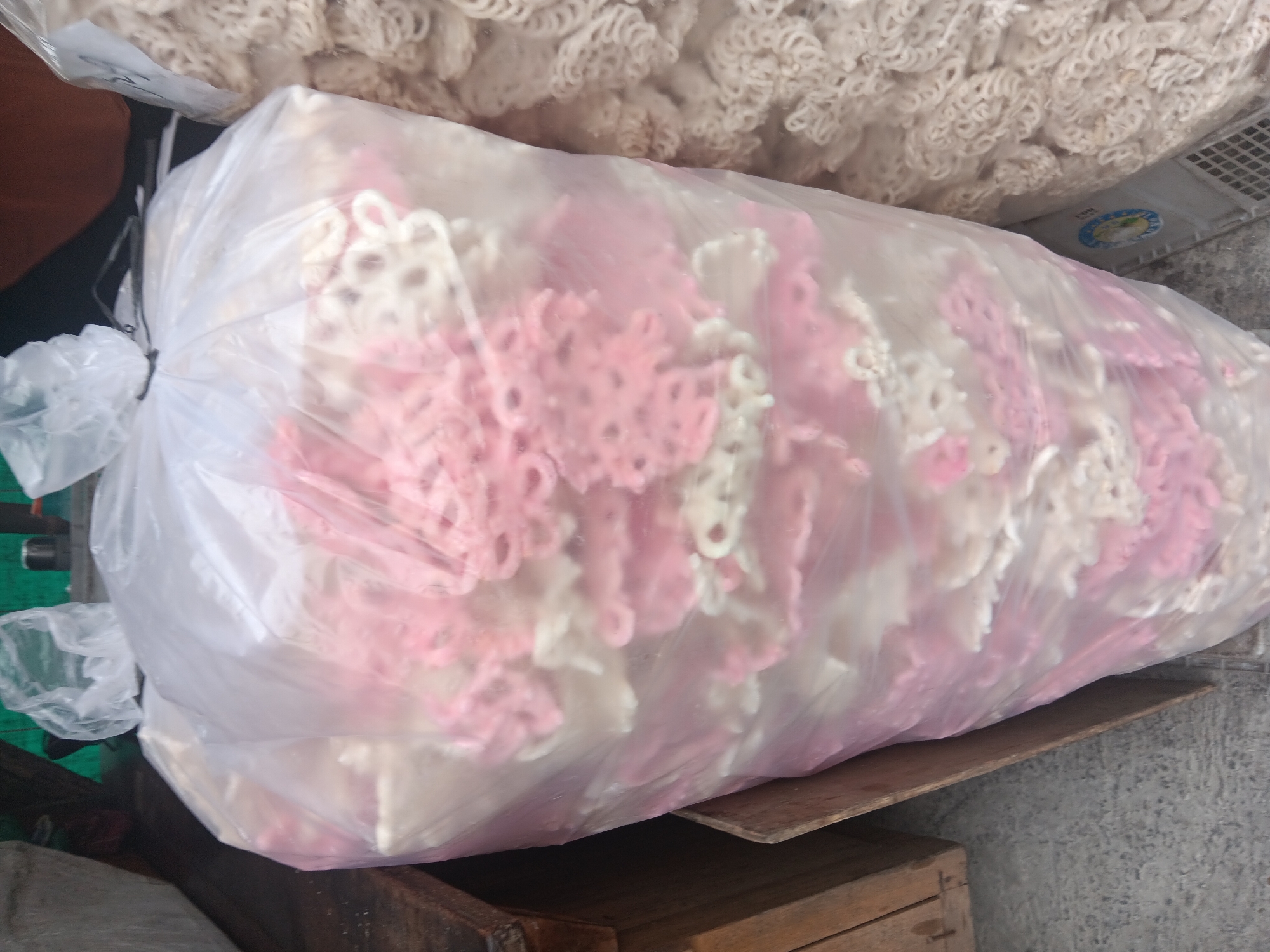 In Batursari, Indonesia, a large, transparent plastic bag is filled with a substantial quantity of pink and white crackers. The crackers, characterized by their intricate, lace-like patterns, are stacked vertically within the bag, creating a dense visual display. To the right of the main bag, another clear plastic bag contains a similar assortment of crackers, but these are exclusively white and exhibit a more coiled, circular form. The bags are positioned on a concrete surface, with a wooden crate or shelf visible on the left side of the frame. The overall scene suggests a retail or market environment where these food items are being stored or sold.