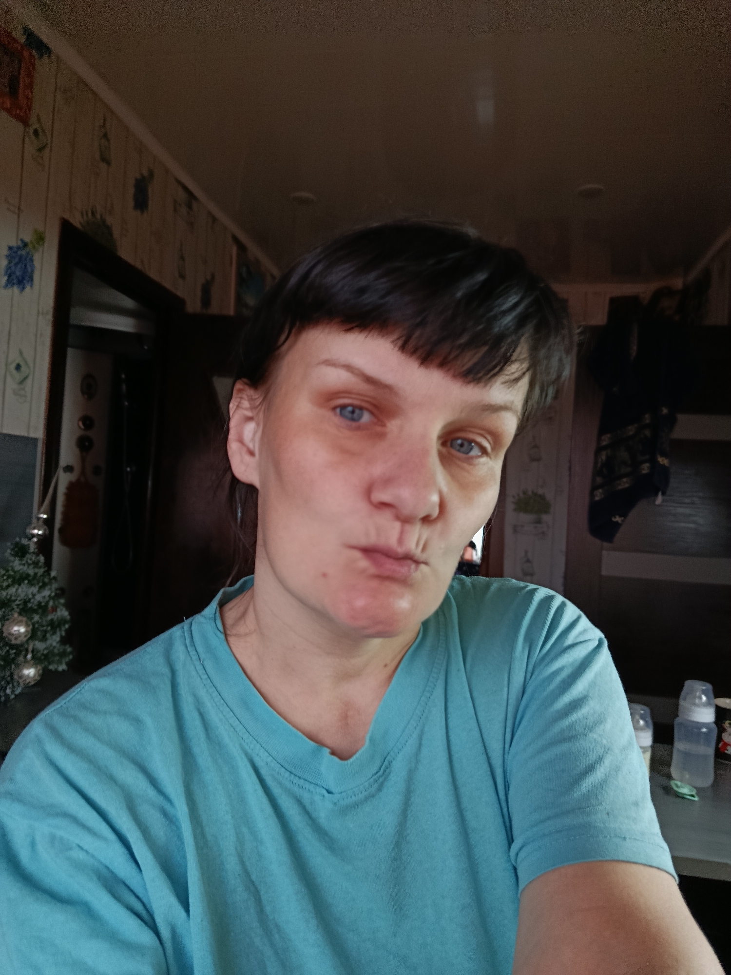 A medium-close-up, slightly low-angle indoor portrait captures a woman with dark, short hair and bangs, and blue eyes. She is looking directly at the camera with a playful, pouting expression, her lips puckered. Her complexion shows some redness, particularly around her eyes. She is wearing a plain, light teal or turquoise short-sleeved t-shirt.
The setting appears to be a domestic interior, likely a home in Фоки, Russia. To the left of the woman, a small, artificial Christmas tree adorned with silver ornaments is visible, suggesting the time of year is late autumn or winter. Behind her, the walls are covered with wallpaper featuring a patterned design, predominantly in muted beige and brown tones with occasional dark blue or green accents. A dark doorway is visible in the background on both the left and right sides of the frame.
On the right side of the image, beyond the woman's shoulder, a shelf or table holds two baby bottles – one appearing full of a clear liquid and another empty – along with a small, decorated cup or mug. A dark, patterned fabric item hangs on a wall or door in the upper right background. The lighting is soft and even, characteristic of indoor ambient light, suggesting it could be daytime. No specific activity beyond posing for a selfie is discernible. There is no clearly readable text visible in the image.