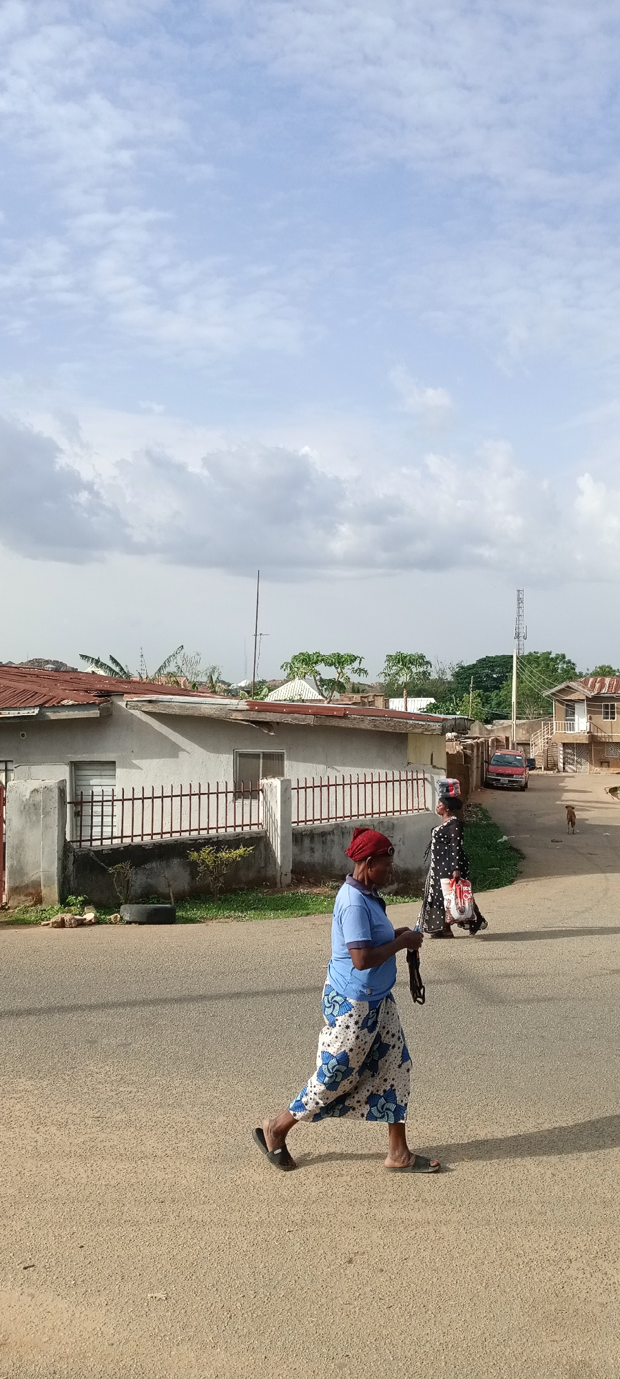 The image depicts a street scene in an urban area. The street is made of dirt or asphalt. Two women are walking along the street. The woman closest to the camera is wearing a blue shirt, a patterned white and blue skirt, a red headscarf, and sandals. She is carrying a dark object in her left hand. Further down the street, another woman is carrying a container on her head and a bag in her hand. A red car is parked in the street in front of a tan colored house with a concrete staircase leading to the front door. A dog is walking in the street. A low, horizontal building with a red tile roof and a metal fence runs along the left side of the street. A cell phone tower is located in the background, behind the houses. The sky is overcast.