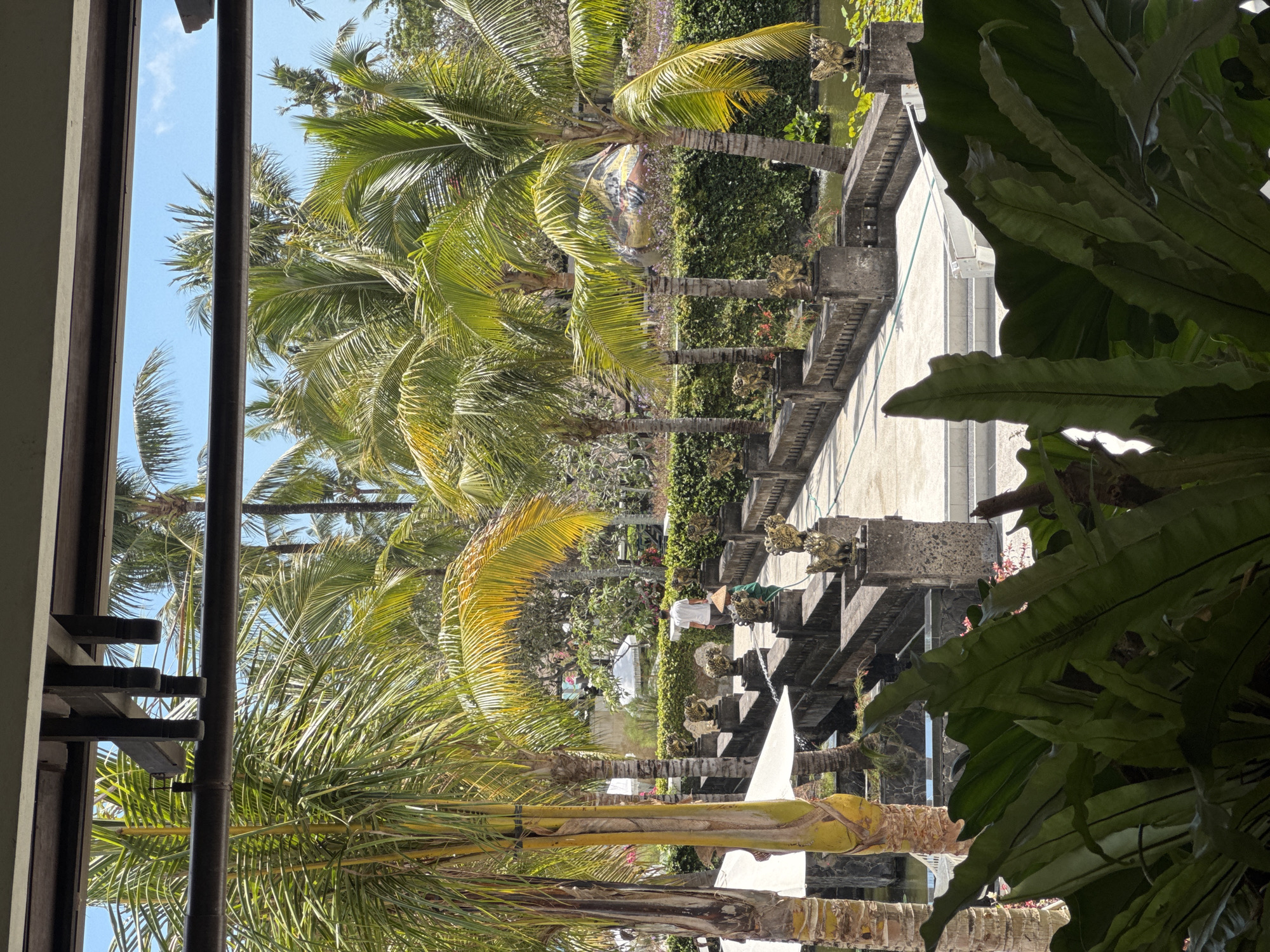 The image depicts a tropical resort environment in Kuta Selatan, Indonesia, featuring lush palm trees and meticulously manicured gardens. In the foreground, large, dark green fern leaves partially obscure the view. Beyond them, a stone bridge with ornate, lion-like sculptures at its pillars spans a body of water. A person wearing a conical hat and a green garment is visible on the bridge, tending to plants with a hose. Another individual in a white shirt walks further along the bridge. The background reveals more palm trees, white umbrellas, and a densely hedged wall, under a clear blue sky. The scene suggests a tranquil, well-maintained outdoor area within a resort.