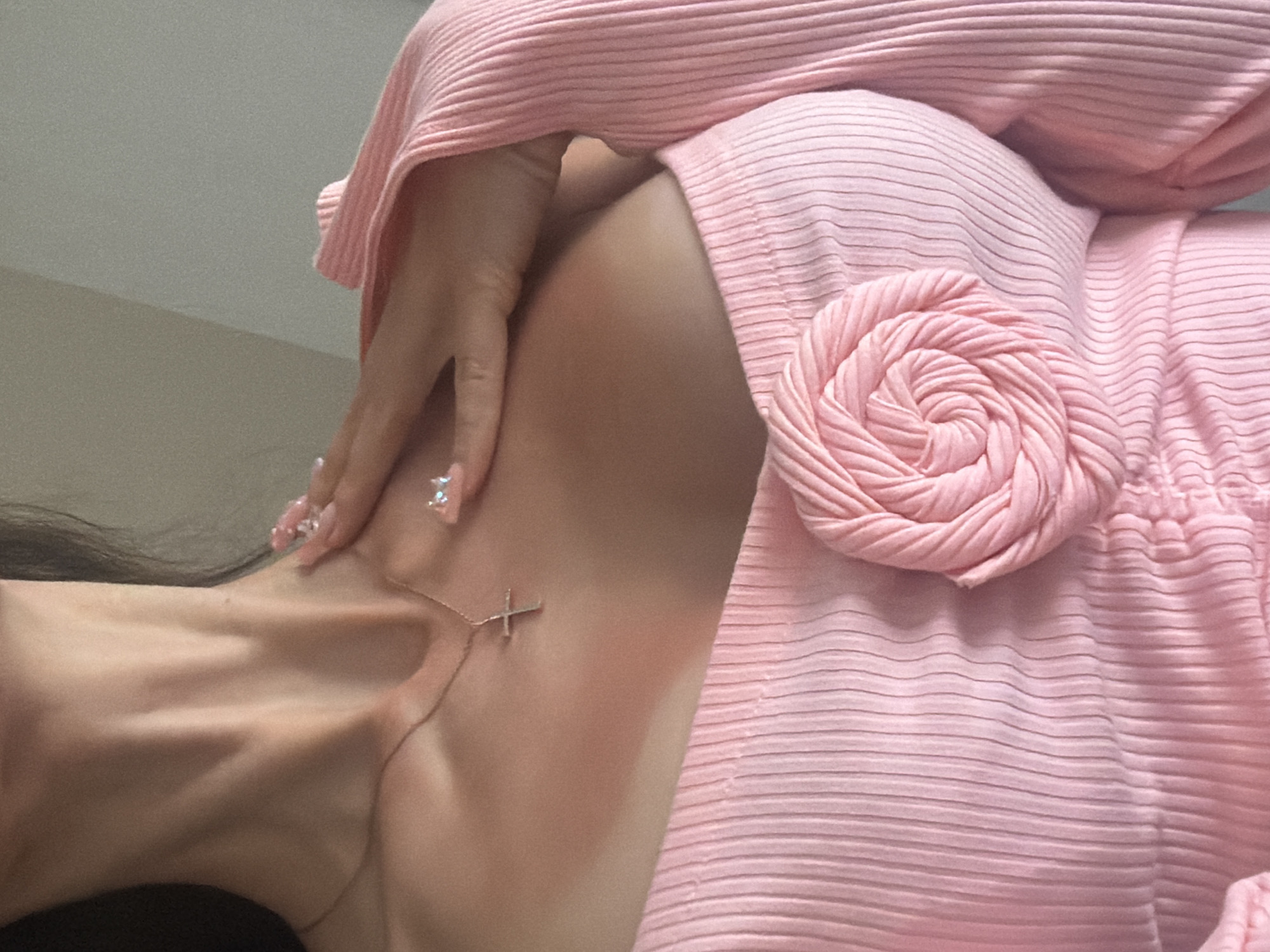 The image is a close-up, chest-up shot of a person wearing a pink, ribbed knit top. The top is off-the-shoulder and features a rose-shaped appliqué on the left side of the chest. The person's right hand, adorned with long, light-colored nails decorated with small gems, is placed on their neck and shoulder. A delicate gold chain with a cross pendant rests on the person's collarbone. Subtle tan lines are visible on the chest and neck area. The background is out of focus, showing a neutral, light-colored wall and a hint of natural light. The image does not provide any information about a specific location in a city or country.