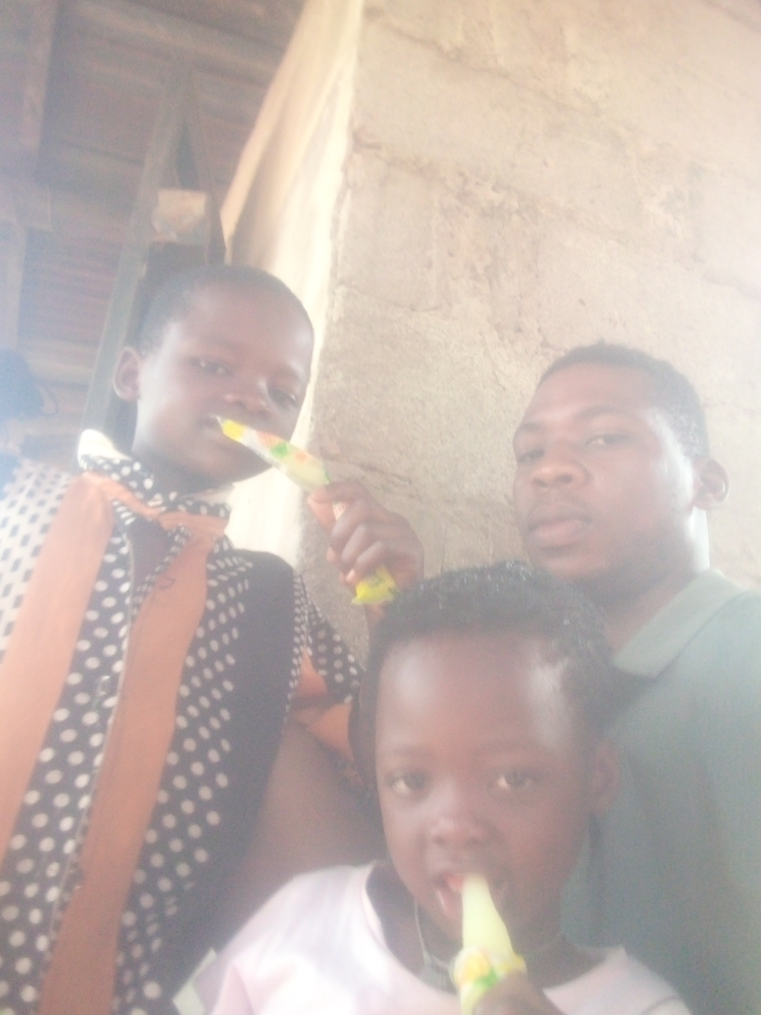 Three individuals are captured in this close-up photograph. At the forefront is a child with dark hair, clad in a light-colored shirt. To the left and slightly behind, another child is seen wearing a patterned shirt with a tied portion at the chest. Behind and to the right, an adult male is present. All three subjects appear to be eating or holding a slender, cylindrical object resembling an ice pop or similar treat. The background is composed of an aged wall. The location appears to be in the city of Unknown, Unknown.