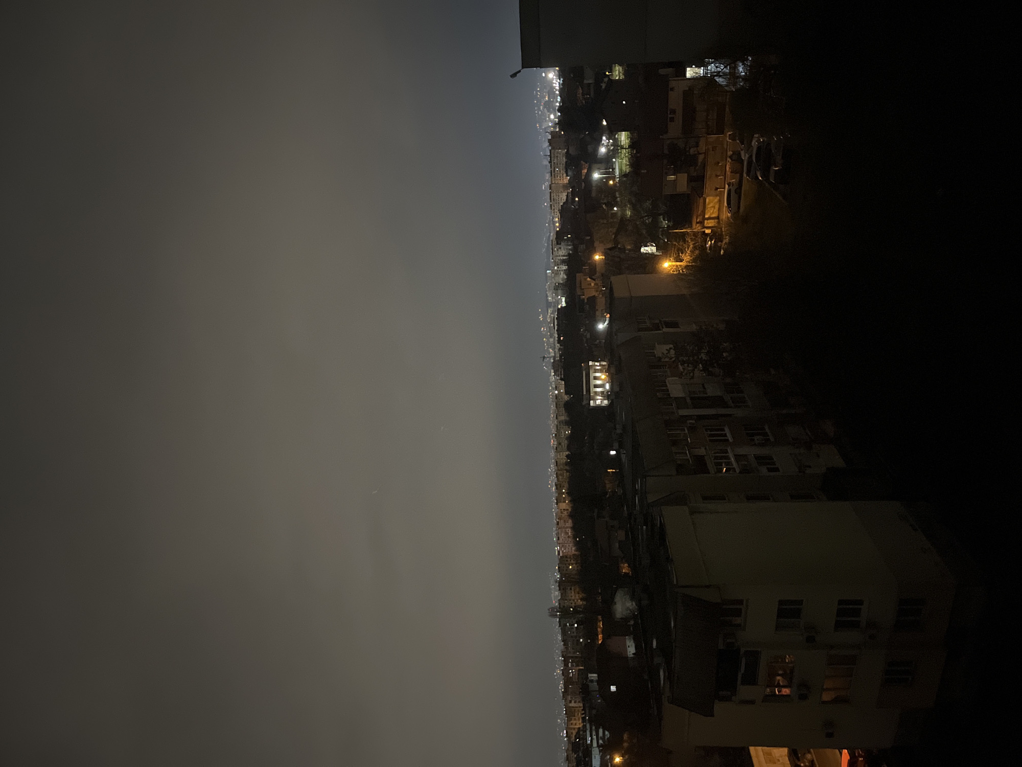 The image depicts an urban nighttime scene, possibly in Sofia, Bulgaria, characterized by a thick haze or fog obscuring the cityscape. The left side of the image shows the hazy sky, devoid of distinct features. On the right, buildings and streetlights are partially visible, their details softened by the fog. The architecture appears to be a mix of mid-rise residential buildings, some with illuminated windows. Streetlights cast a warm glow, creating a contrast with the dim sky. The image lacks any visible people or specific actions. The environment is urban, with an emphasis on the built environment and the atmospheric conditions.