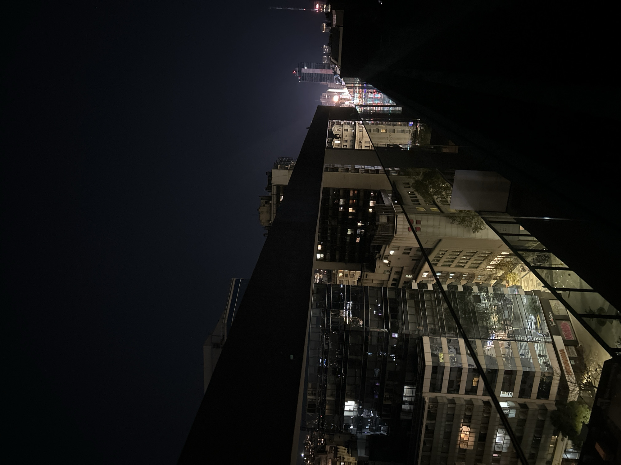 The image depicts a night scene in Buenos Aires, Argentina, specifically a view from a high-rise building. To the left, dark sky fills a large portion of the frame. Dominating the center and right are several buildings, some with illuminated windows suggesting residential or commercial occupancy. A portion of a structure, possibly part of the building from which the photograph was taken, is visible as a dark, angled form obscuring parts of the urban landscape. In the distance, the skyline of Buenos Aires is visible, characterized by clusters of bright lights and tall structures, some appearing to be high-rise buildings. The overall impression is that of a densely populated urban environment.