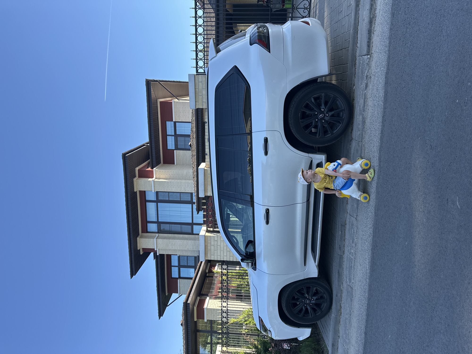 A white SUV is parked on a paved street next to a modern two-story house. A young child wearing a yellow shirt, blue shorts, a patterned hat, and yellow shoes is riding a white toy horse with yellow and blue accents beside the SUV's rear wheel. The house has a tan brick exterior with dark-framed windows. A black metal fence runs along the property line. A contrail is visible in the clear blue sky. The location is possibly within the city of Nizhny Novgorod, Russia.