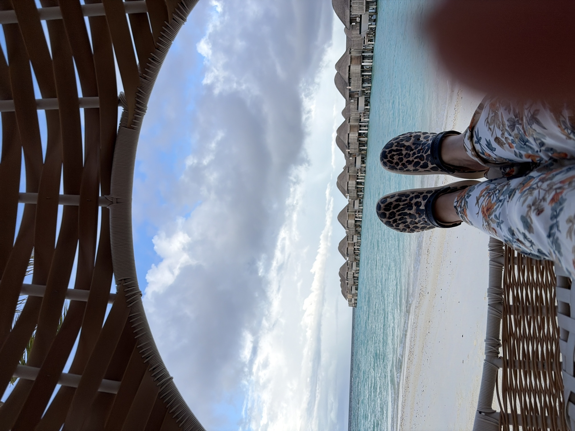 The image shows a first-person perspective of a person's legs and feet, clad in floral print pants and leopard-print clogs, resting in a woven chair. The view is framed by a curved, wooden canopy structure. Beyond the chair, a white sand beach meets the turquoise waters of the Indian Ocean. In the distance, a row of overwater bungalows with thatched roofs stretches across the horizon. The sky is partly cloudy, with patches of blue visible. The scene is set in Halaveli, Maldives, a tropical island known for its luxurious resorts and pristine natural beauty.