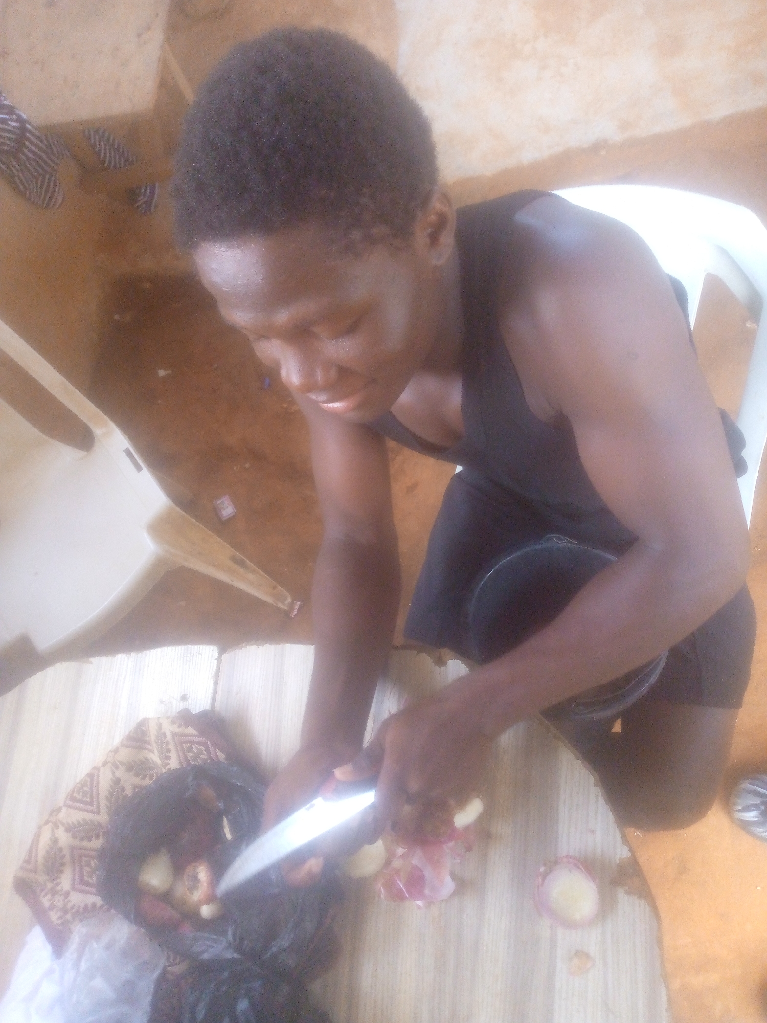 This image captures a person, likely male, with dark skin and short, dark, curly hair, engaged in food preparation. They are wearing a black sleeveless top and dark shorts, and are in a squatting or kneeling position, looking down at their task.
The central activity involves the person using a silver knife to peel or cut light-colored vegetables, which appear to be onions, judging by the reddish-purple peels scattered on a light-colored surface. Some unpeeled vegetables are visible in a black plastic bag that rests on a patterned fabric mat or cloth.
The setting appears to be an indoor or covered outdoor space during the daytime, indicated by the bright, diffused lighting. The immediate ground where the person is working is a light-colored surface, possibly a mat or wooden planks. In the background, two white plastic chairs are partially visible, and the floor or wall further back has a light brown or orange hue. The person shows a focused expression, possibly with a slight smile, indicating concentration on their work. There is no visible text in the image.