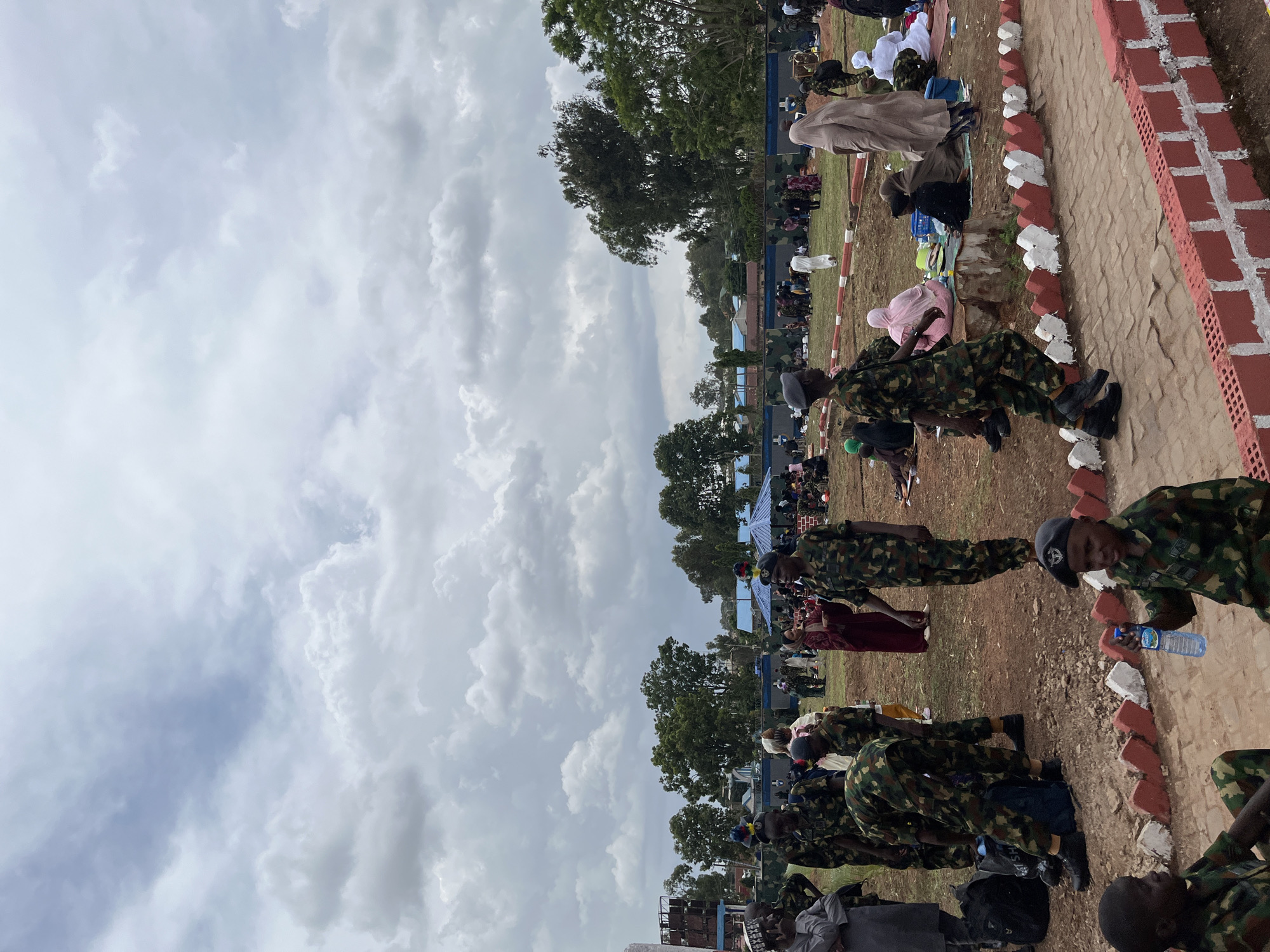 In Barakin Lerawi, Nigeria, a large outdoor gathering is underway, characterized by a mix of military personnel and civilians. The scene unfolds under a heavily clouded sky, suggesting an overcast day. In the foreground, several individuals in camouflage military uniforms are visible. Some are standing, while others are seated on the ground. A young soldier in the lower right corner holds a bottle of water.
Further back, more people are dispersed across a grassy area, many seated on mats or the ground. Some civilians are dressed in traditional attire, including women in hijabs and abayas. Military personnel appear to be present in a supporting or security role, mingling with the civilian population.
Structures in the background include buildings with blue roofs, tents, and a wall adorned with a camouflage pattern, separating the gathering area from what appears to be more open terrain. Trees dot the landscape behind the buildings, indicating a semi-urban or rural setting. The overall impression is one of a community event or a gathering related to a military presence, with people congregating outdoors.