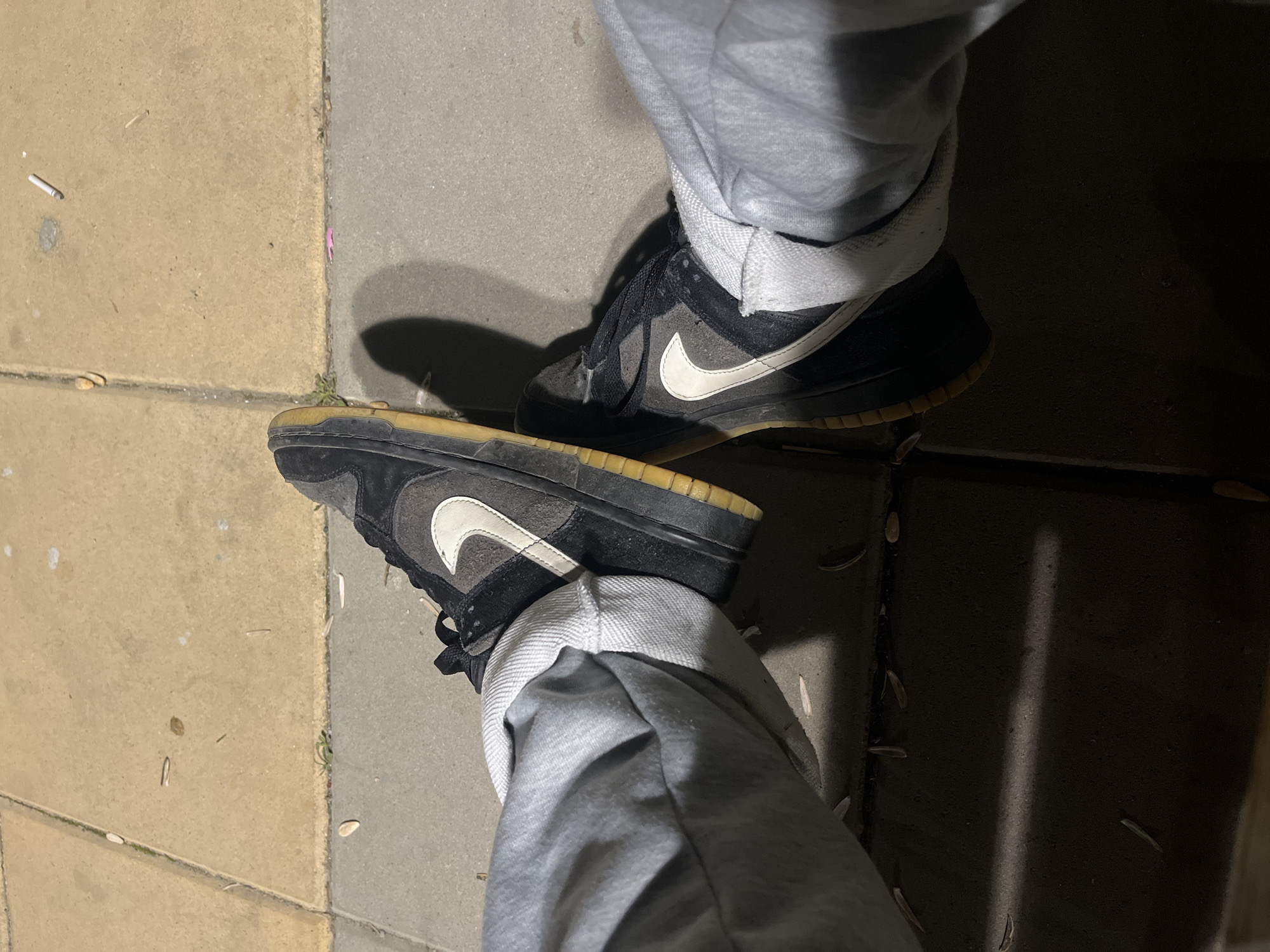 The image shows a person's legs and feet, wearing gray sweatpants and black Nike Dunk Low sneakers with white swooshes and yellowed soles. The person is standing on a paved surface, composed of beige and gray concrete slabs. Debris, possibly seeds or plant matter, is scattered on the ground. The lighting suggests it is either dusk or a dimly lit area. The image does not contain sufficient information to determine if the photo was taken in Lozarevo, Bulgaria.
