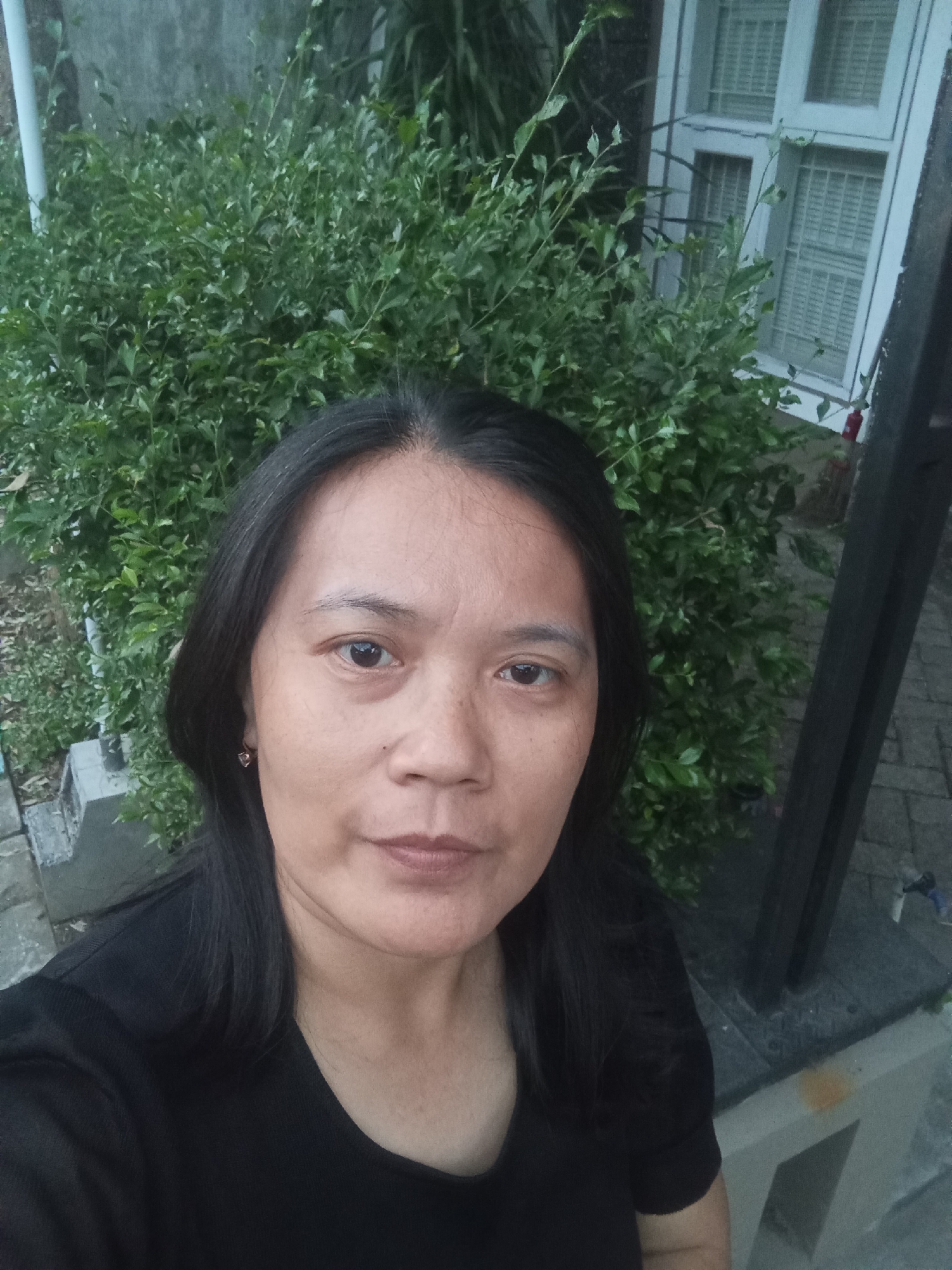 This is a selfie taken outdoors in Malang, Indonesia, by a woman with long, dark hair and a black shirt. She is standing in front of a dense green hedge and a building with white-framed windows. The ground appears to be paved with stone.