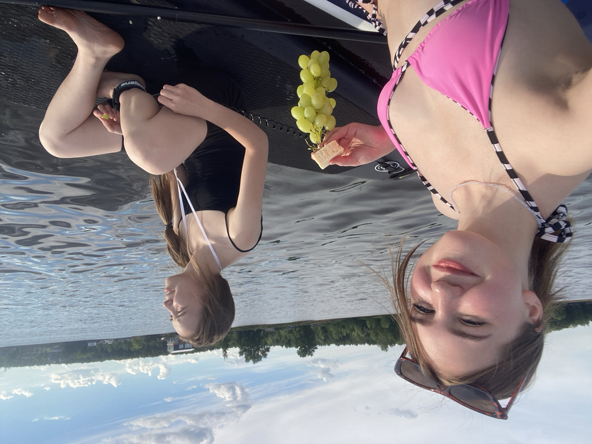 Two young women are on a paddleboard on a body of water, likely a lake or river, with a treeline and buildings visible on the distant shore. The woman in the foreground is facing the camera, smiling, and wearing a pink bikini top and sunglasses. She holds a bunch of green grapes with a piece of food in her hand. The woman in the background is facing away from the camera, looking out at the water, and is wearing a black swimsuit. The water is rippled, and the sky is partly cloudy. The scene appears to be set in Chaikovsky, Russia, given the context of the location.