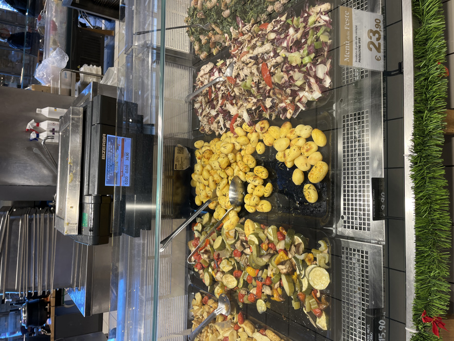 The media file captures an indoor food display counter, likely within a supermarket or deli in Rozzano, Italy. Various prepared food items are showcased in transparent containers behind a glass barrier, suggesting a self-service or assisted-service buffet.
The display features four distinct dishes:
* The topmost container, partially visible, appears to hold a mixed greens or vegetable salad.
* Below it, a full container holds "Insalata di pollo alla senape" (chicken salad with mustard), containing chopped chicken, radicchio, and other ingredients. This dish is priced at €23.90, as indicated by a sign also reading "Menú delle Feste" (Holiday Menu) and "PRODUZIONE IPER".
* The middle container is filled with golden-brown roasted potatoes, seasoned with herbs like rosemary. A partially visible sign indicates "Patate al Forno" (Baked Potatoes) for €15.90.
* The bottom container contains mixed roasted vegetables, including zucchini, bell peppers, and possibly onions, also priced at €15.90 for "Miste" (mixed).
Serving spoons are placed in the potato and mixed vegetable containers. To the left of the food display, a Bizerba brand weighing scale is visible. Its digital display shows "0.020 0.020 34.90", indicating a weight of 0.020 units and a price of €34.90, likely per kilogram. The background shows stainless steel surfaces, suggesting a commercial kitchen or food preparation area. A green, artificial garland runs along the right side of the display, hinting at a festive or holiday season.