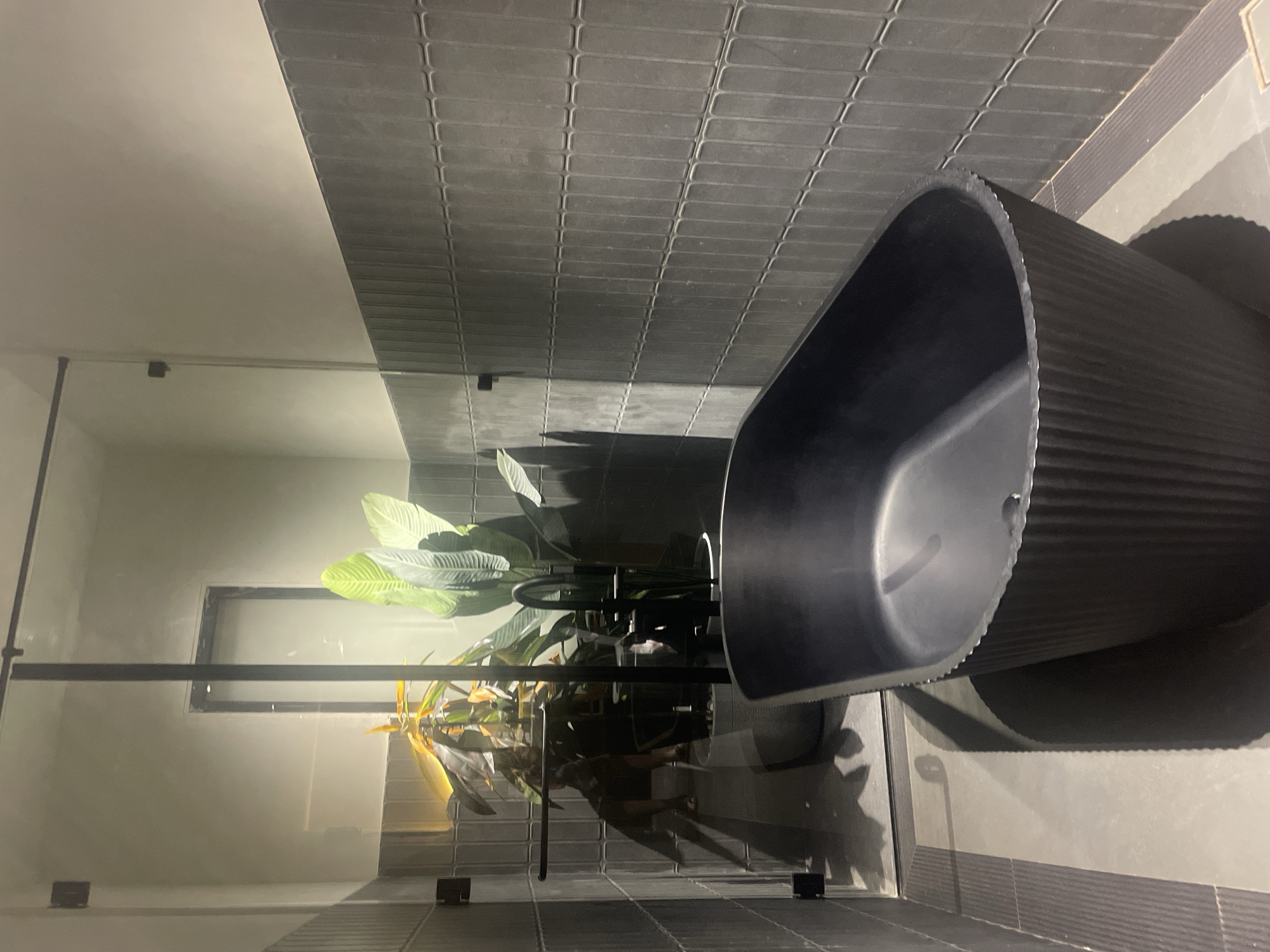 The image shows a modern bathroom. The walls are covered in dark gray, rectangular tiles. A sleek, black bathtub with ribbed detailing sits prominently in the space. A potted plant with large, green leaves is positioned behind the tub, adding a touch of nature to the setting. The scene is well-lit, and the floor appears to be made of similar dark tiles as the walls. The location is Chopdem, India.