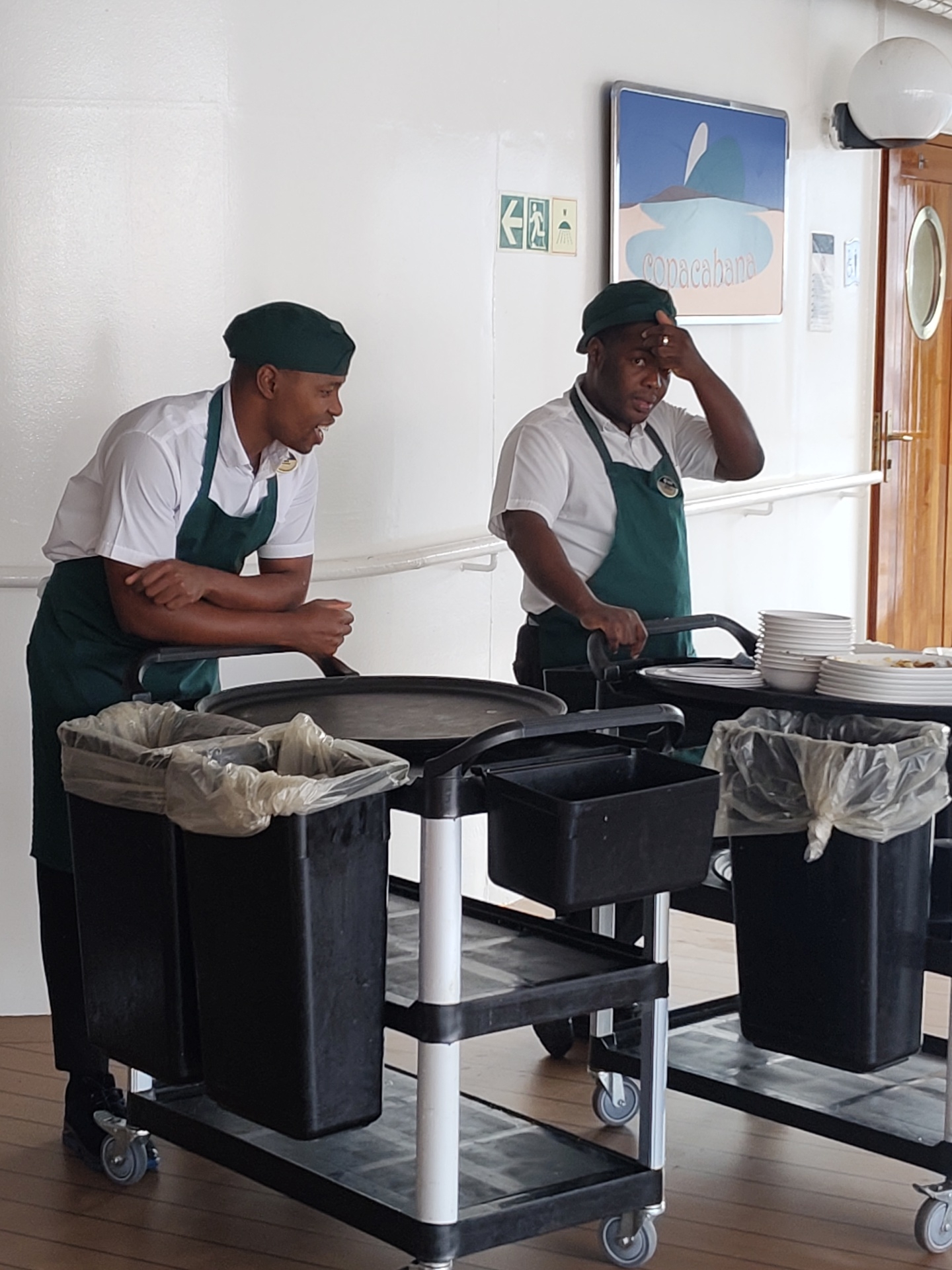 The image shows two men wearing dark green caps and aprons over white shirts, standing next to service carts. The man on the left leans on his cart and is speaking, while the man on the right has his hand on his forehead. Each cart has refuse containers. Behind them is a white wall with a door, a light fixture, and a picture of "Copacabana" with what appears to be Sugar Loaf Mountain. A green exit sign is visible. It's likely the men are cruise ship workers. The location cannot be determined as Genoa, Italy without further information.