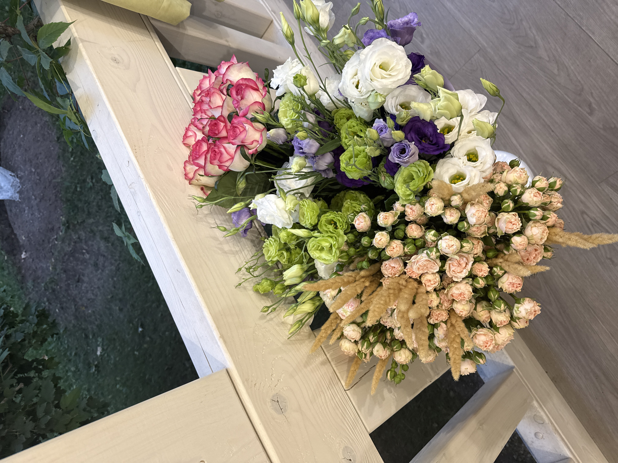 A bouquet of flowers is arranged in a white vase on a wooden floor. The bouquet is comprised of various flowers including pink roses with white edges, green flowers, purple flowers, and small pink roses. There are also dried grass accents within the bouquet. The bouquet is positioned on a wooden deck with a light-colored railing. In the background, a green, leafy area with dirt is visible. The scene appears to be outdoors, possibly on a balcony or porch, in Arinj, Armenia.