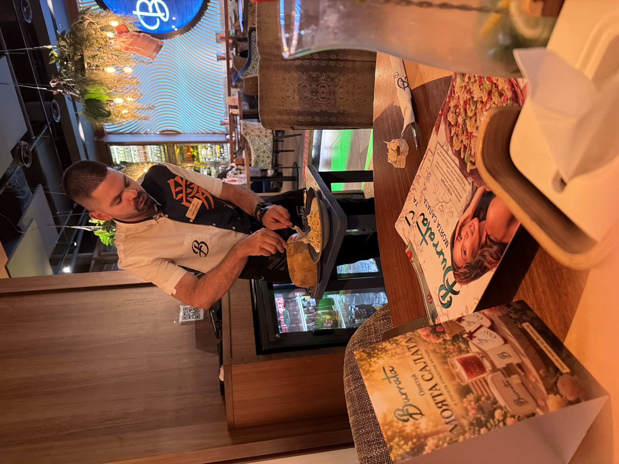 The image depicts an indoor scene at the Burrata Italiana restaurant in Sofia, Bulgaria. In the center, a male server, identified by his name tag as Konstantin Ivanov, is actively preparing food at a small, dark serving table. He wears a white short-sleeved shirt with a black and orange patterned vest featuring the Japanese character "愛" (Ai - love) and the restaurant's "B" logo. He is using a knife to cut into a large, round, yellowish food item, likely a wheel of cheese or a bread bowl, from which he is serving pasta or risotto onto a dark plate.
In the immediate foreground, a wooden dining table holds various items. Two "Burrata" menus are visible: one standing upright displaying "МОЯТА САЛАТА" (My Salad) with imagery of a candle and gift boxes, and another lying flat featuring a woman and a salad with pomegranate. Also on the table are a white paper napkin bearing the "B" logo, a small crumpled piece of paper, a white tissue dispenser, and a clear glass pitcher containing a beverage with ice and citrus slices.
The background reveals the restaurant's interior. Wooden wall paneling is on the left. Hanging decorative plants with embedded lights adorn the ceiling. A prominent circular blue sign with a large stylized "B" logo and a wavy textured background is illuminated on the back wall. Further back, various dining tables and chairs with patterned upholstery are visible. Behind the server, a mini-refrigerator stocked with drinks is present, and a shelf above it displays a QR code. A strip of green lighting is visible along the floor in the deeper background.