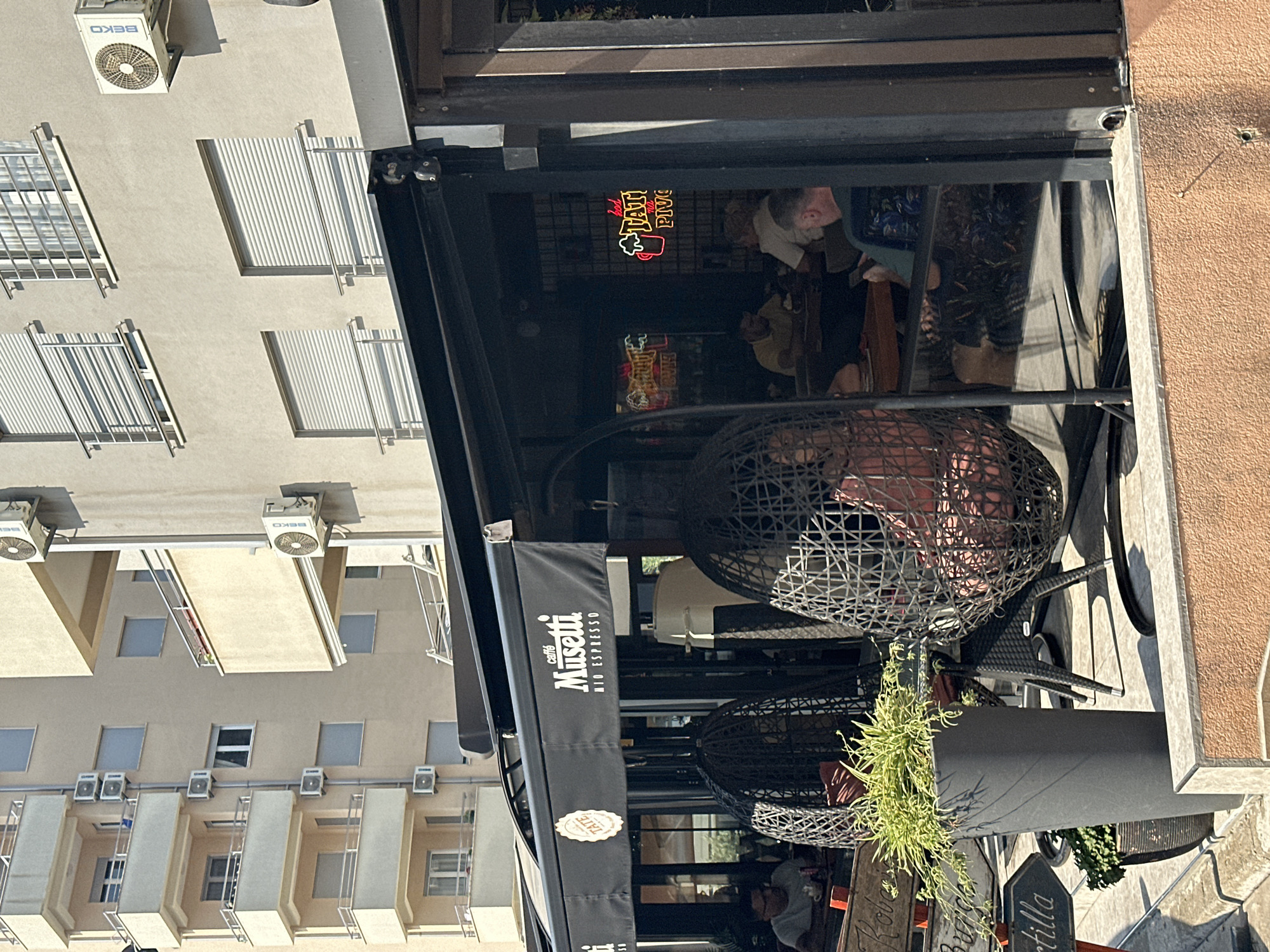 This image depicts the outdoor seating area of a cafe called "Caffè Musetti MID ESPRESSO" in Budva, Montenegro, on a sunny day. The cafe has a black awning with the cafe's logo. In the foreground, there are two large, black, woven hanging chairs with pink cushions. A tall, dark gray planter with lush green foliage is positioned to the left of the chairs. Beyond the chairs, inside the cafe, several patrons are seated at tables. One man in the lower left is looking at his phone. Another man, in the middle of the image, is wearing a yellow shirt and appears to be engaged in conversation. A woman with blonde hair is seated at a table further back. A neon sign with a beer mug and the text "TATI PIVO" is visible inside the cafe, suggesting it is also a bar or serves beer. In the background, the facade of a modern apartment building is visible, with balconies and multiple air conditioning units. The overall scene is one of a relaxed, urban cafe environment.