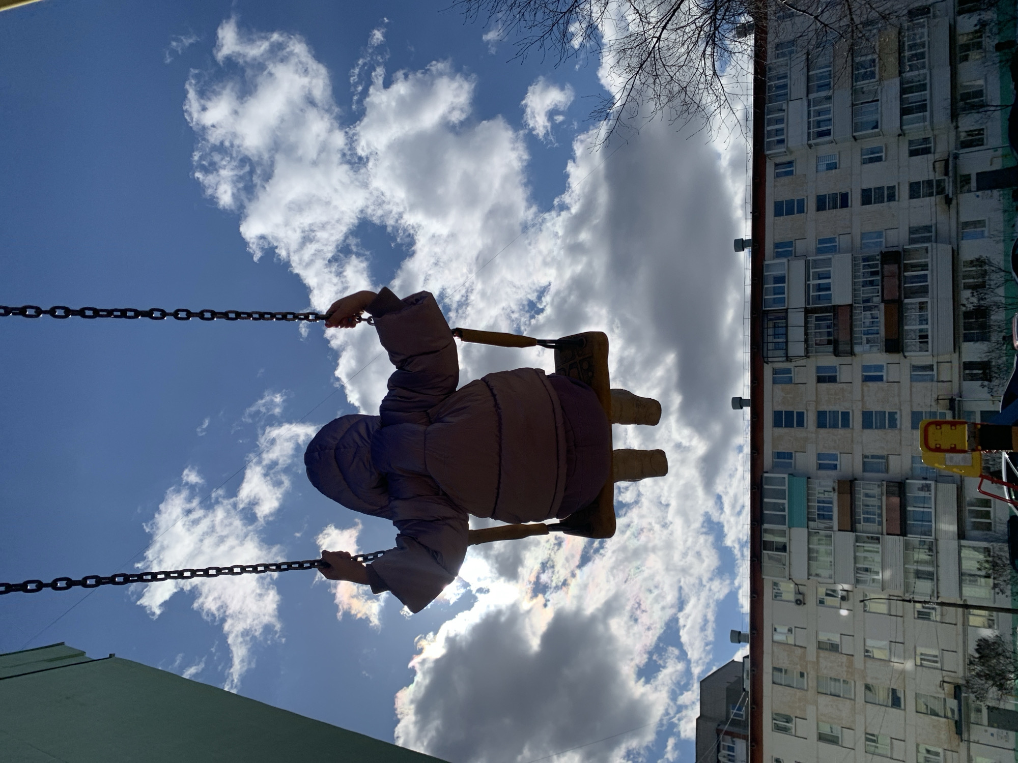 A child in a hooded puffer jacket swings against a backdrop of a partly cloudy, bright blue sky. The child is facing away from the viewer. The swing is suspended by chains. In the background, there is a tall, aging apartment building with numerous windows. There are also some bare tree branches visible. The location is Chaikovsky, Russia.