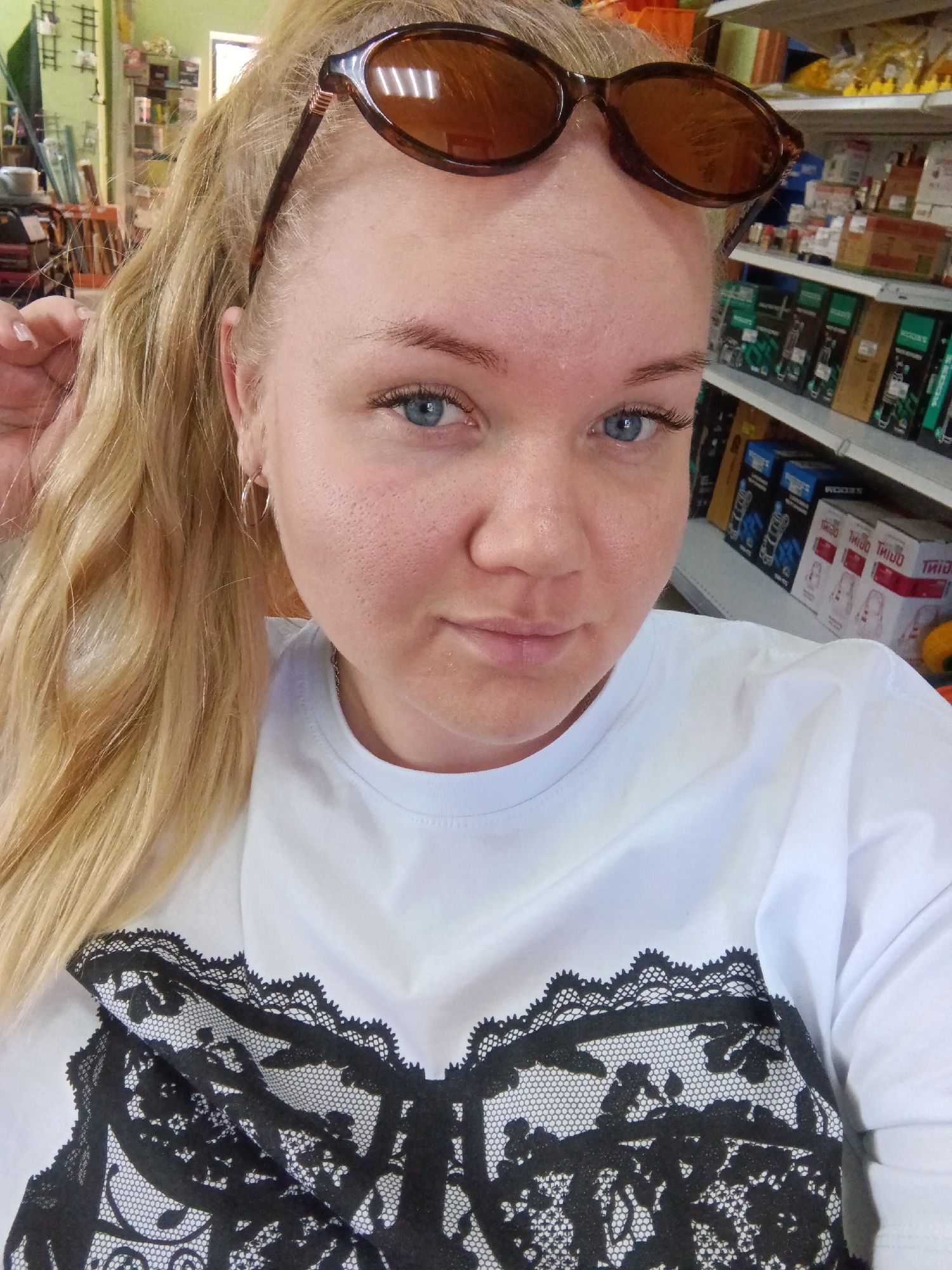 A fair-skinned woman with blonde hair in a ponytail and blue eyes poses for a selfie indoors. She is wearing sunglasses atop her head and a white t-shirt with a black lace design on the front. She has earrings in her ears.
In the background are shelves stocked with various items, suggestive of a store setting. It is impossible to identify the location in the city of Фоки, Russia from the provided image.