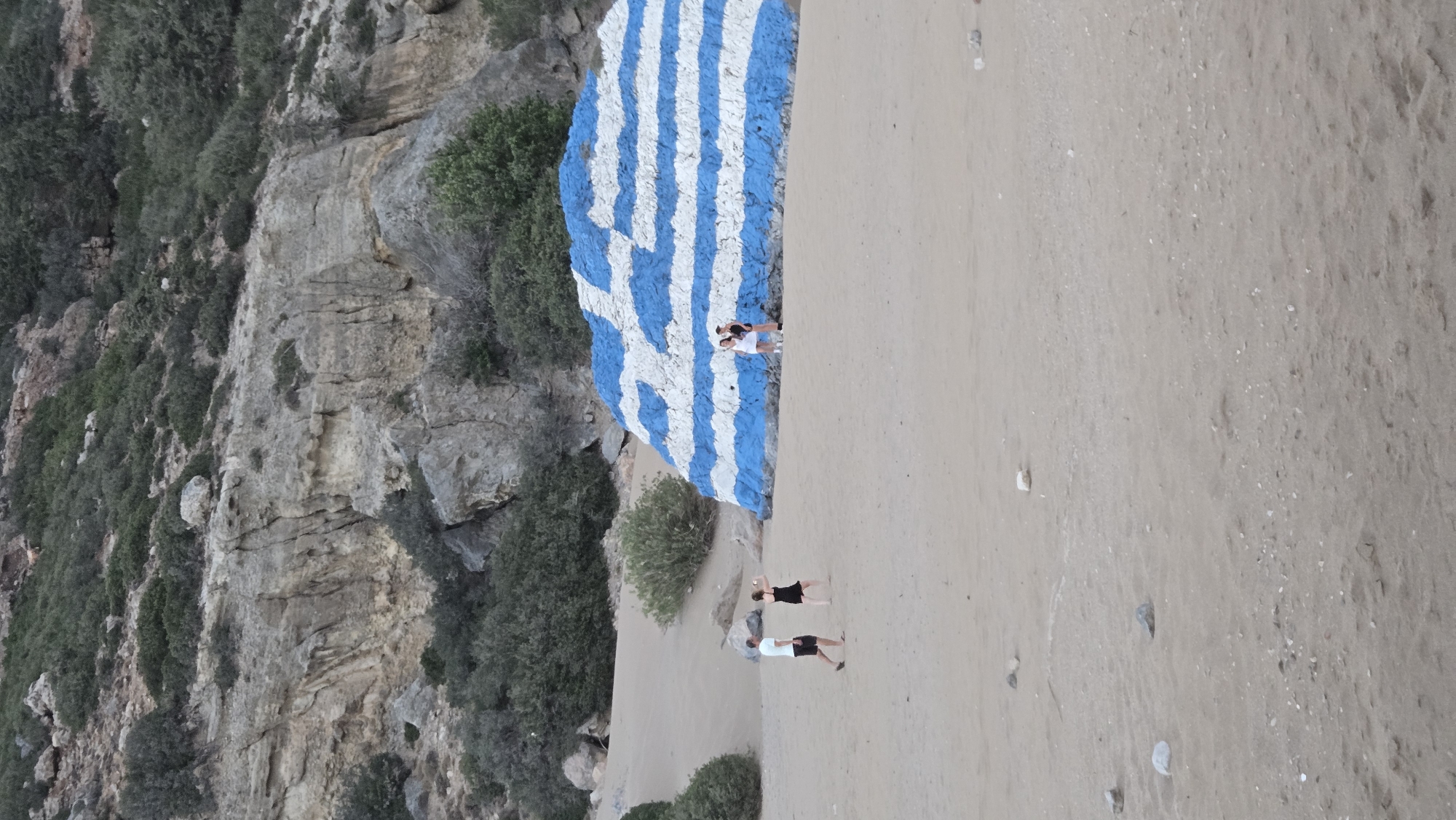 In Kolymbia, Greece, a large rock painted to resemble the Greek flag stands prominently on a sandy beach. The flag design consists of alternating blue and white stripes, with a blue square in the upper left corner containing a white cross. Several people are visible in the scene. To the left of the rock, a man wearing a white t-shirt and dark shorts walks away from the camera. Near him, a woman in a black dress is taking a photo with her phone. To the right of the flag rock, two women are standing, one in a white dress and the other in a dark outfit, seemingly posing for a picture or observing the rock. The background features a rocky hillside covered with sparse vegetation and trees. The beach itself is sandy, with some scattered rocks and pebbles. The overall environment suggests a coastal or beach setting.