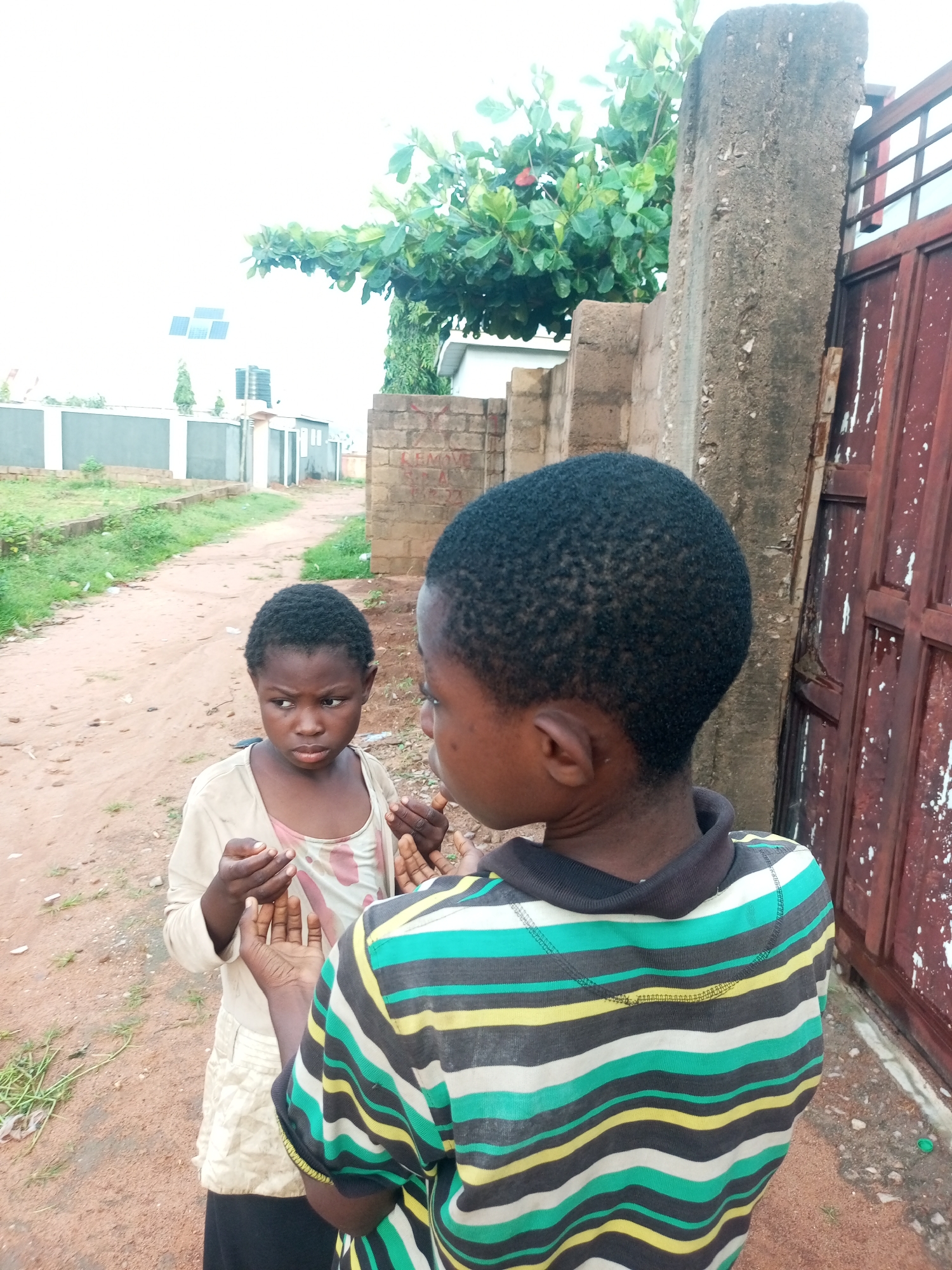 Two young children are standing on a dirt path next to a concrete wall and a red metal gate. The child on the left is facing left and looking down at their hands, which are held up. They are wearing a light-colored shirt with a patterned under-layer and a black skirt. The child on the right is facing away from the camera, looking at the child on the left. They are wearing a striped shirt with black, green, yellow, and white stripes. In the background, there are buildings, a water tank, solar panels, and trees. The overall environment appears to be an outdoor area in a residential or semi-rural part of Tempure, Nigeria.